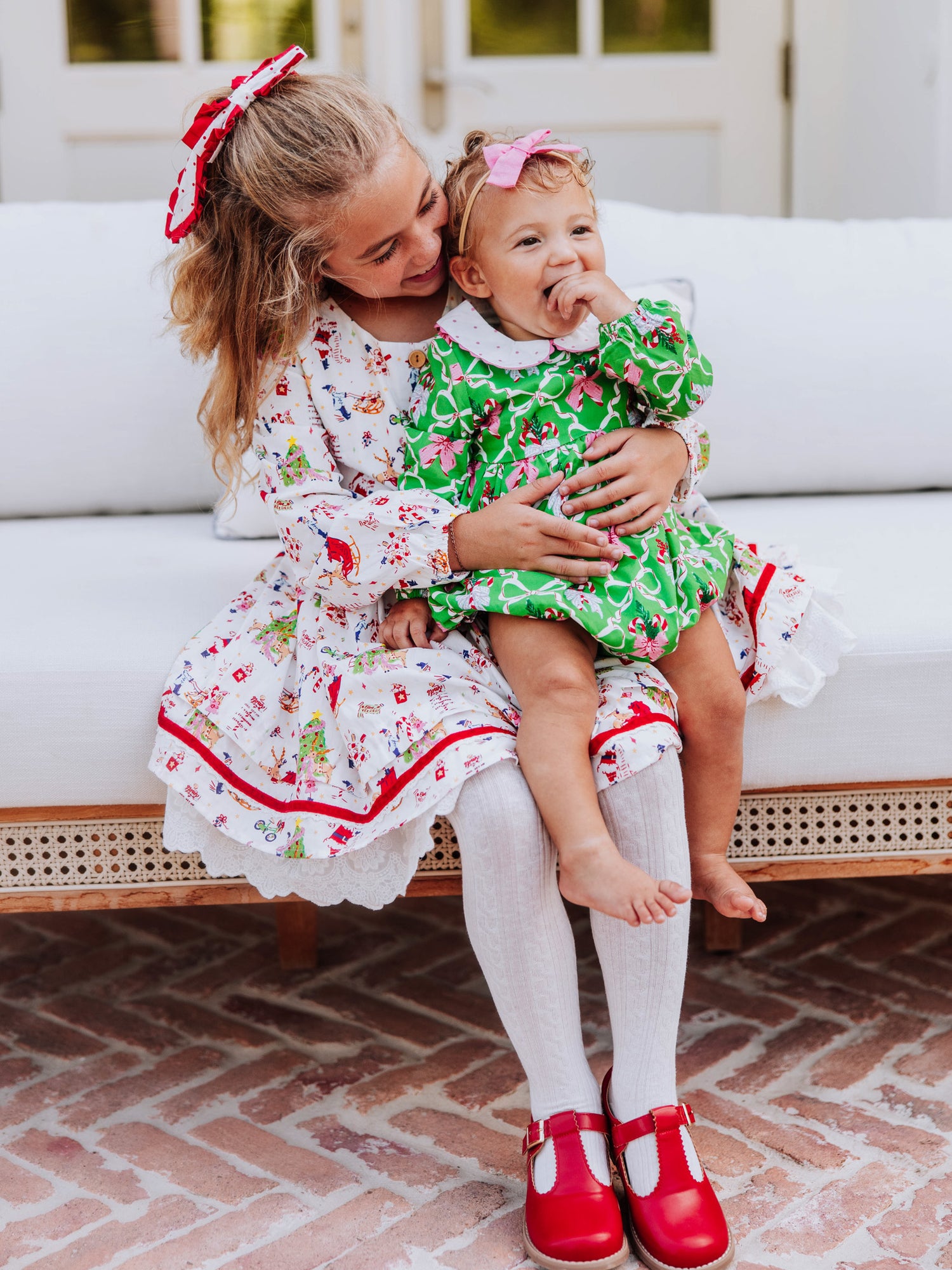 Bigger girl holding a smaller girl and both are sitting on couch. Bigger girl is wearing a holiday themed dress with white tights and red mary janes. Baby is wearing a green holiday themed classic bubble.