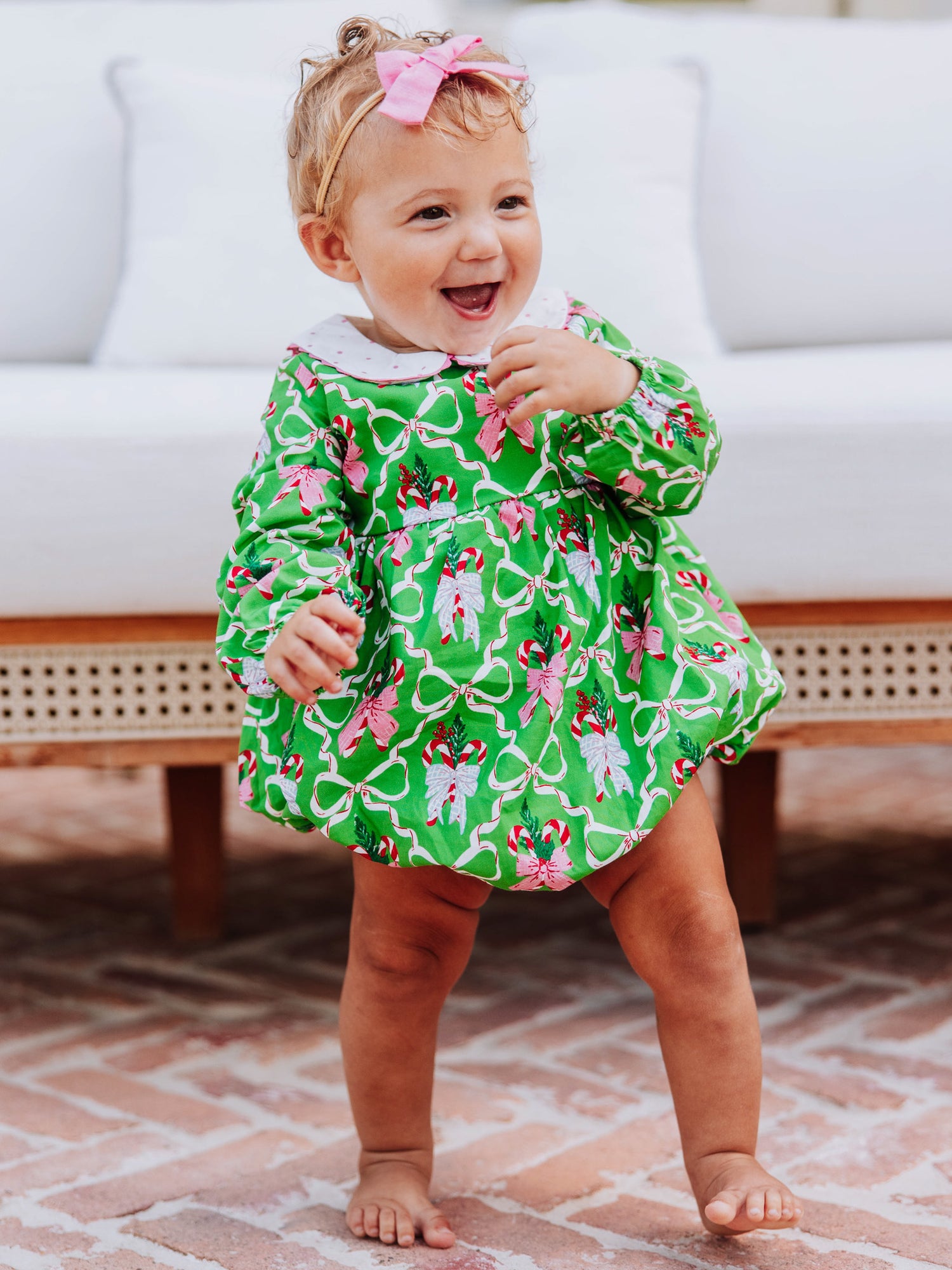 Baby girl standing outside in front of couch wearing a green holiday themed classic bubble.