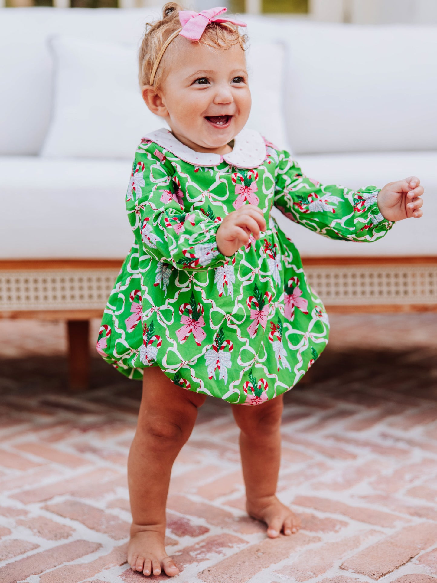 Baby girl standing outside in front of couch wearing a green holiday themed classic bubble.