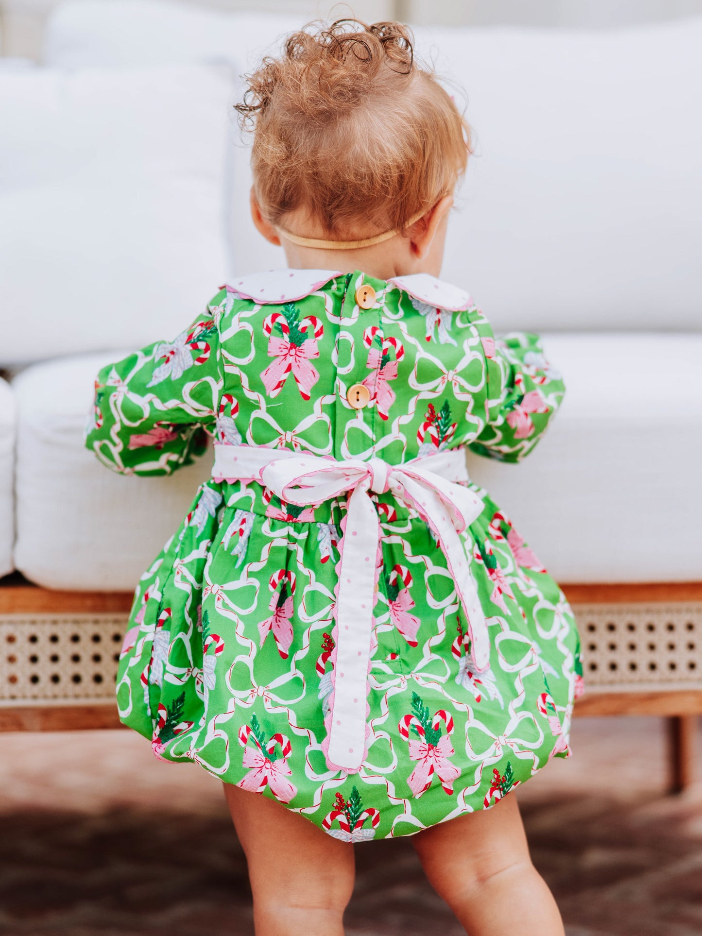 Baby girl standing outside in front of couch wearing a green holiday themed classic bubble. Pic shows the tie at back and the wooden buttons. 