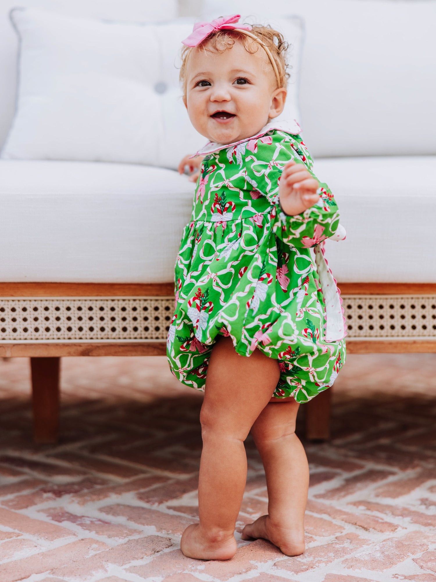 Baby girl standing outside in front of couch wearing a green holiday themed classic bubble.