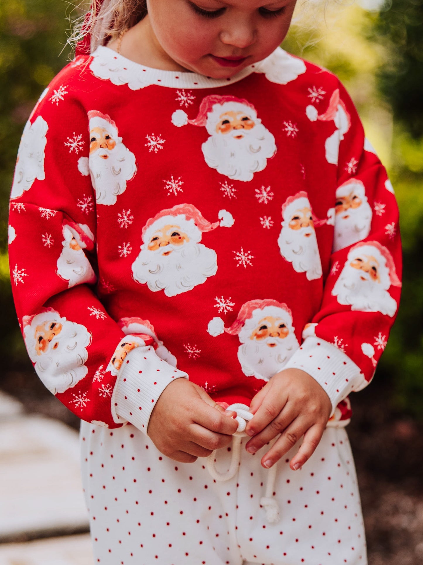 Girl outside wearing a jolly Santa sweat set. Model is tying the drawstring to her bottoms. 