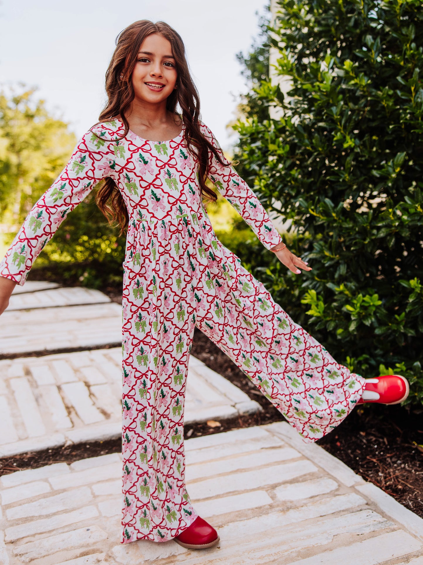 Model standing outside with arms out wearing a holiday bow printed leggy romper and red mary janes