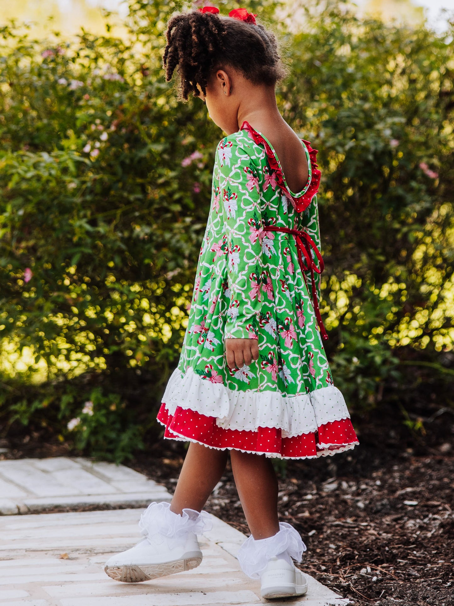 Girl is standing outside holding arms out. She is wearing a holiday themed dress with ruffly socks and white shoes. 