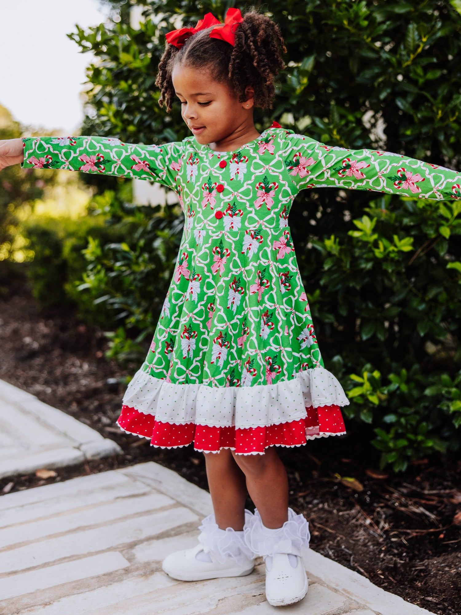 Girl is standing outside holding arms out. She is wearing a holiday themed dress with ruffly socks and white shoes. 
