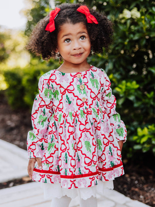Girl standing outside next to greenery wearing a holiday themed dress with bows all over it. 