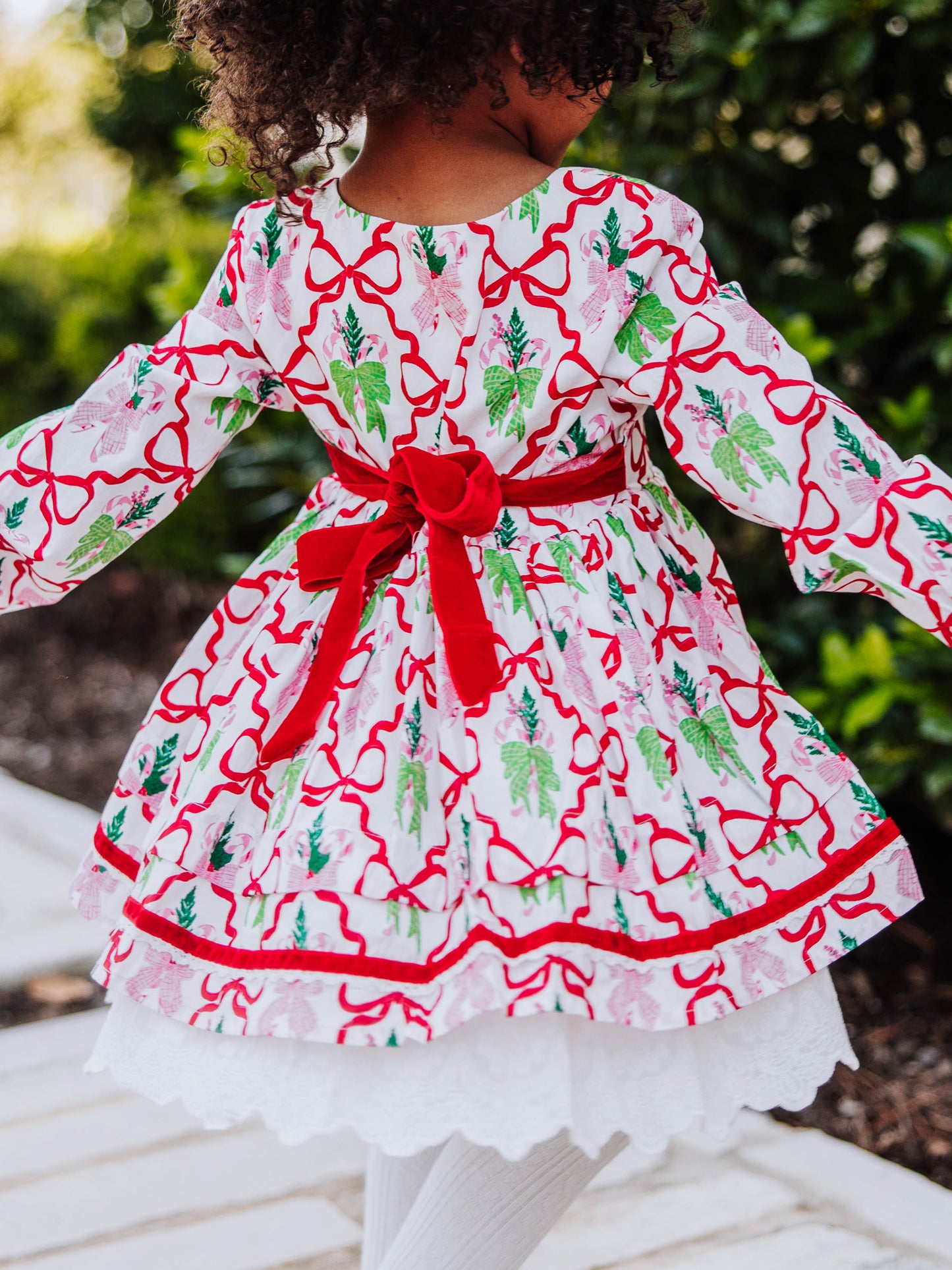 Girl standing outside next to greenery wearing a holiday themed dress with bows all over it. Model is spinning slightly and the back of the dress is shown. Back has a red velvet tie. 