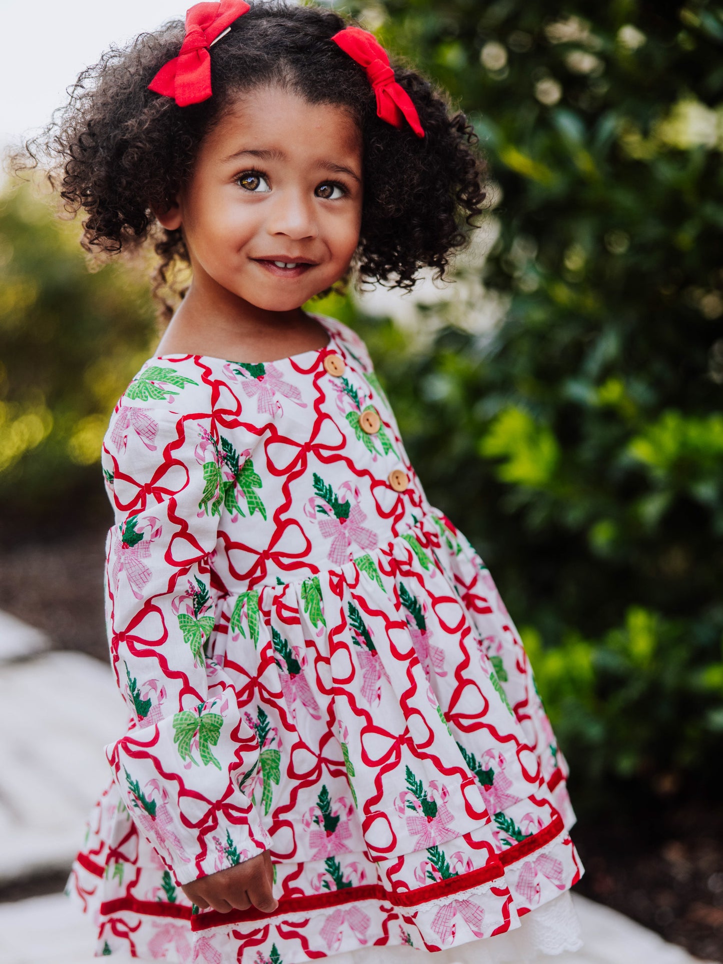 Girl standing outside next to greenery wearing a holiday themed dress with bows all over it. 