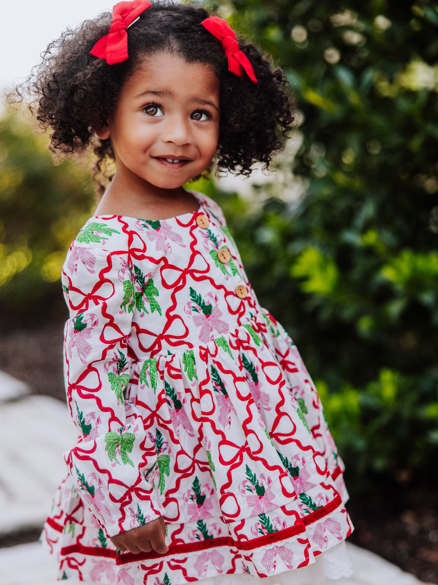 Girl standing outside next to greenery wearing a holiday themed dress with bows all over it. 