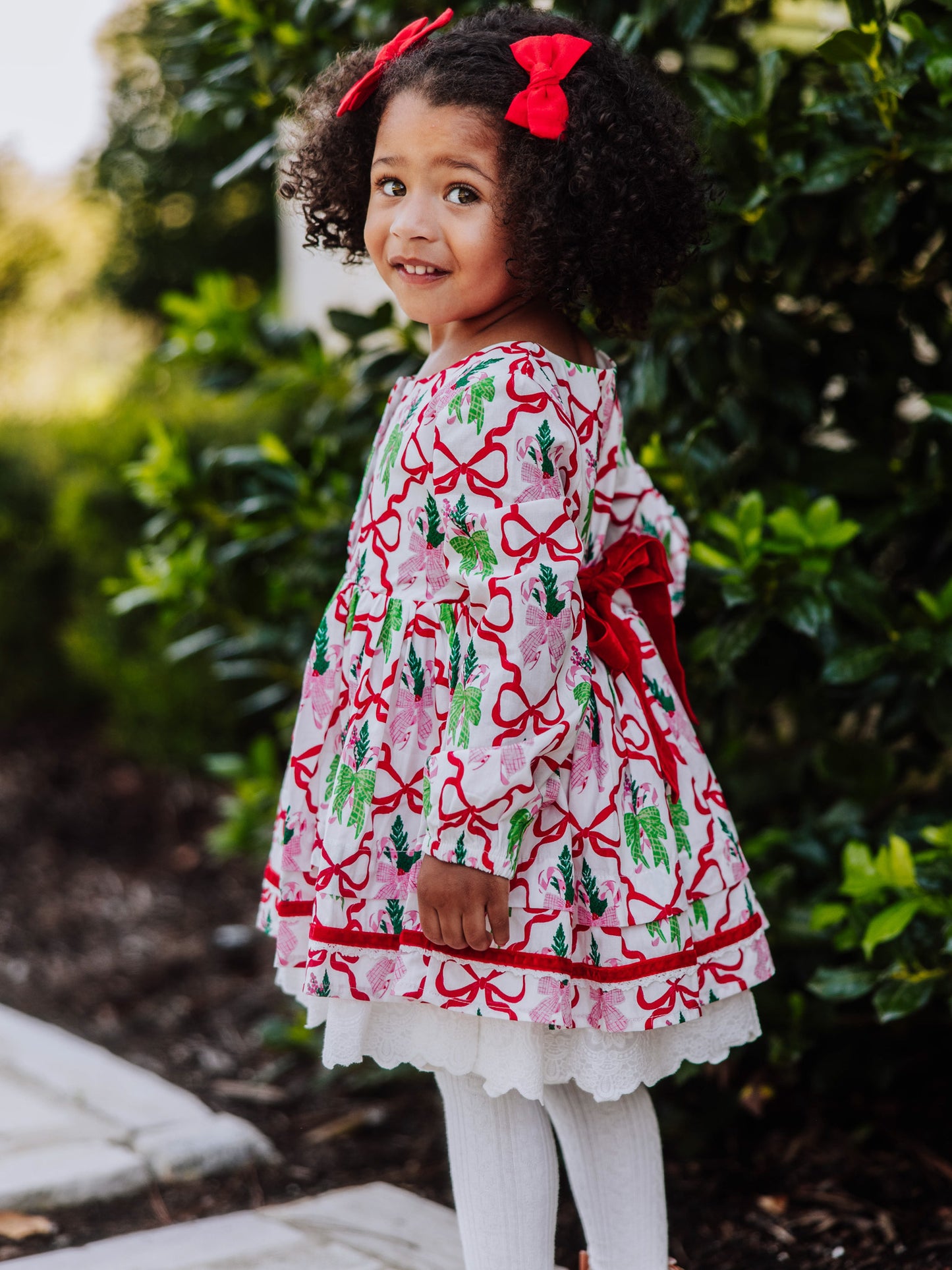 Girl standing outside next to greenery wearing a holiday themed dress with bows all over it. Model is slightly looking over shoulder