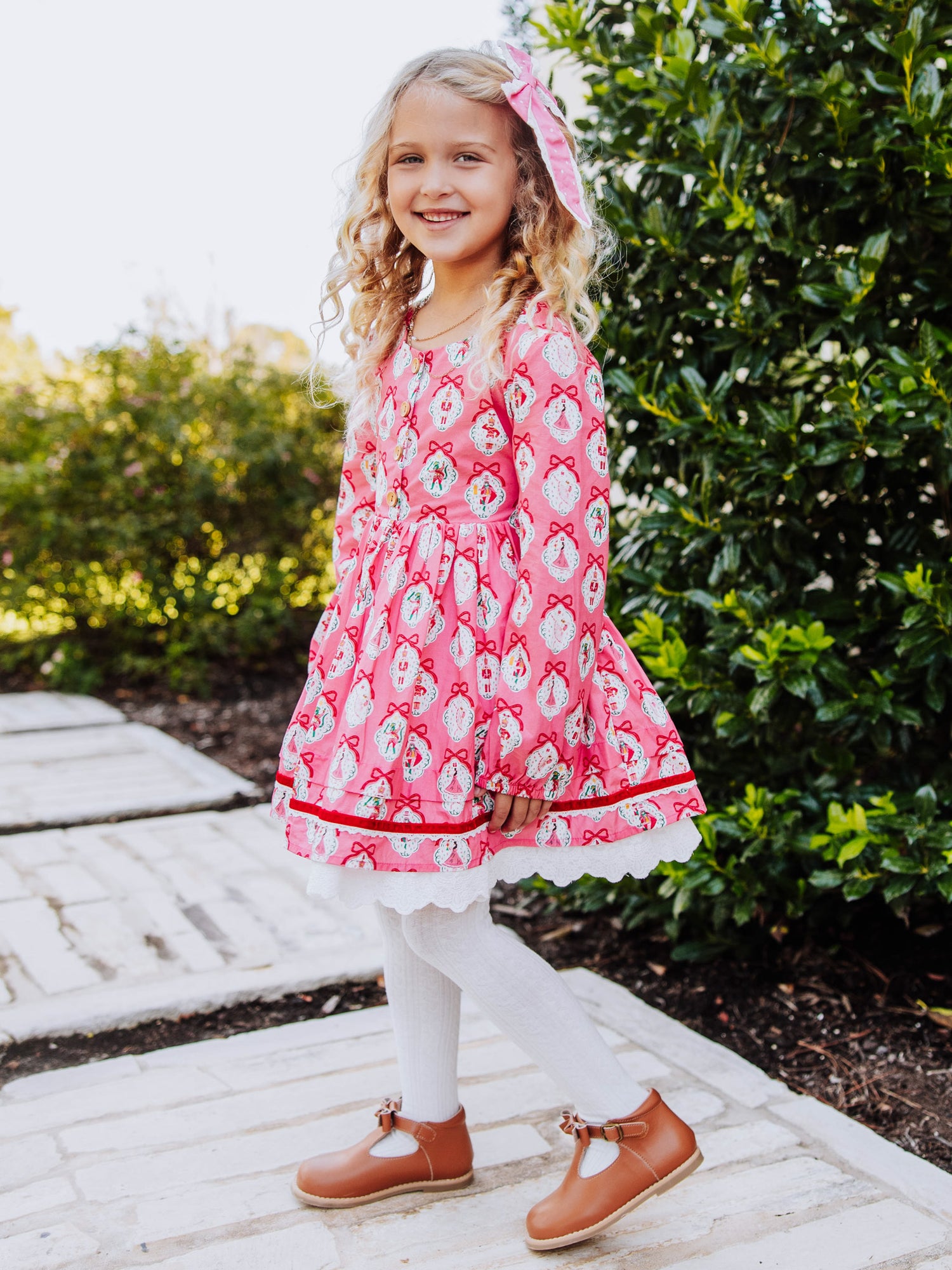 Girl standing outside next to greenery wearing a pink holiday themed dress.