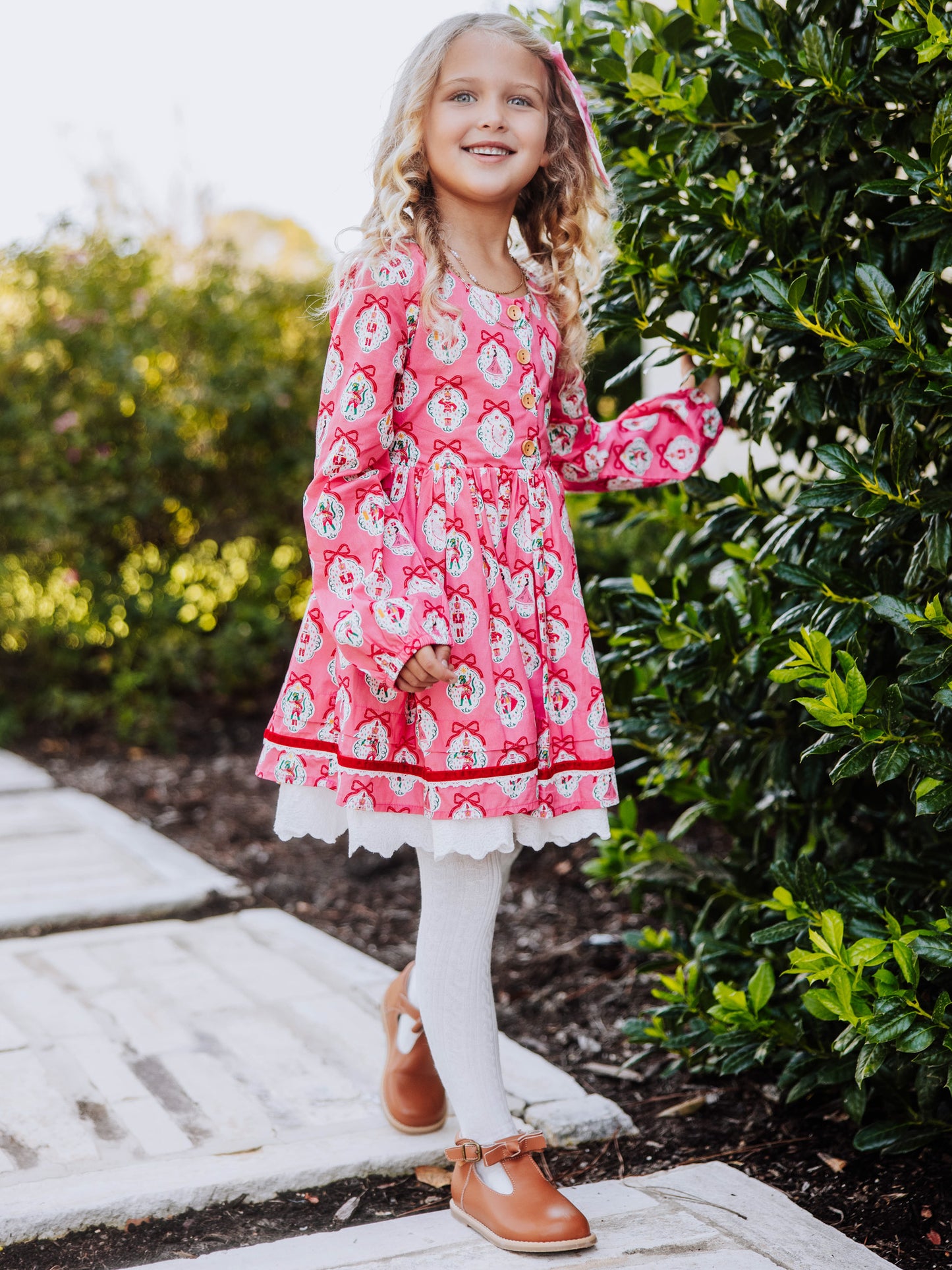 Girl standing outside next to greenery wearing a pink holiday themed dress.