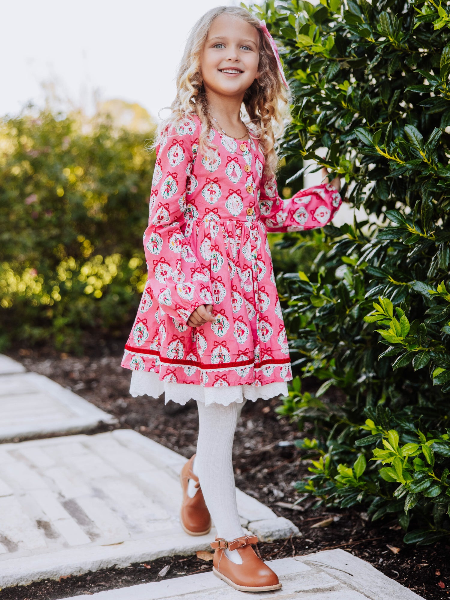 Girl standing outside next to greenery wearing a pink holiday themed dress.