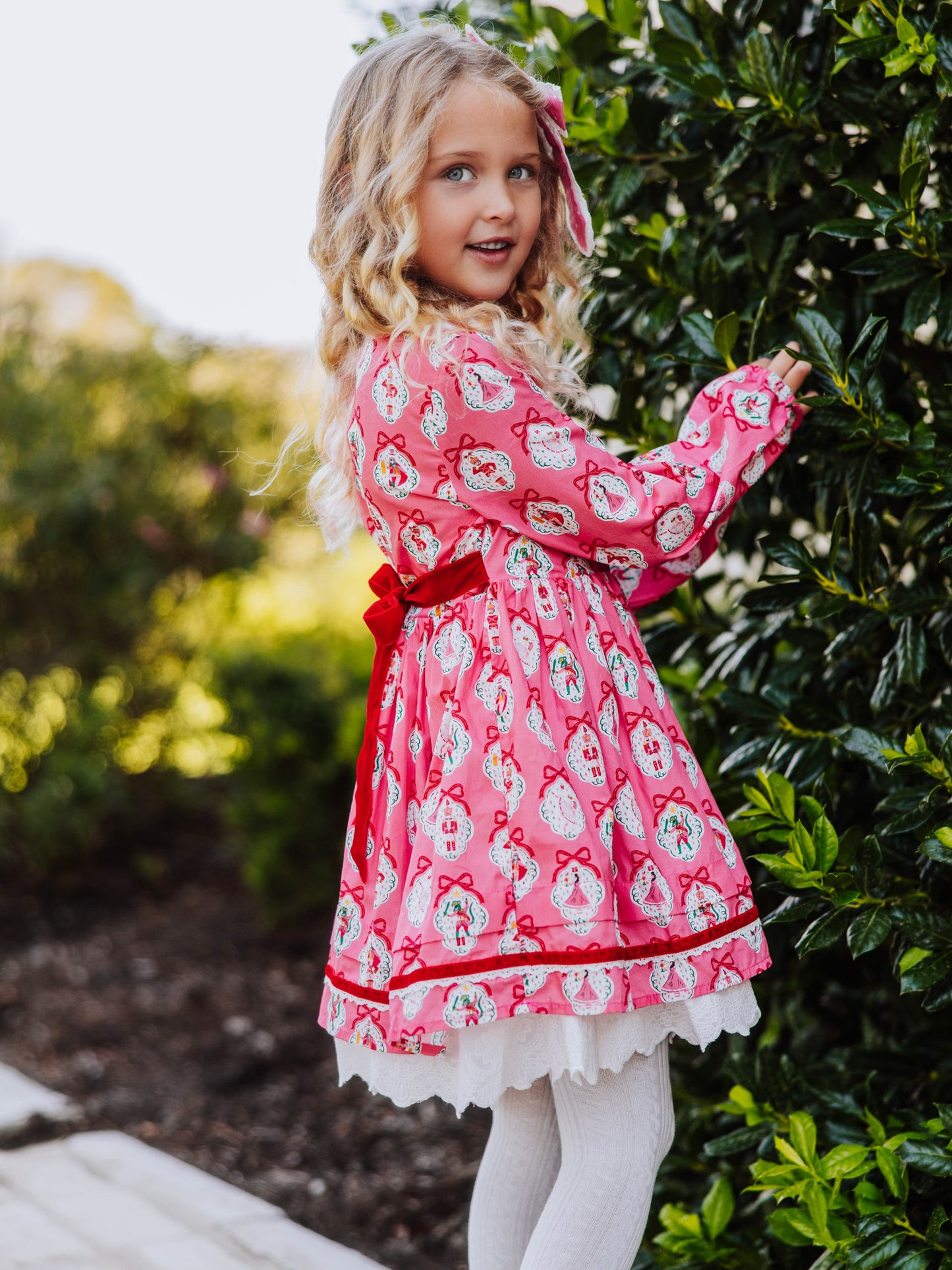 Girl standing outside next to greenery wearing a pink holiday themed dress.