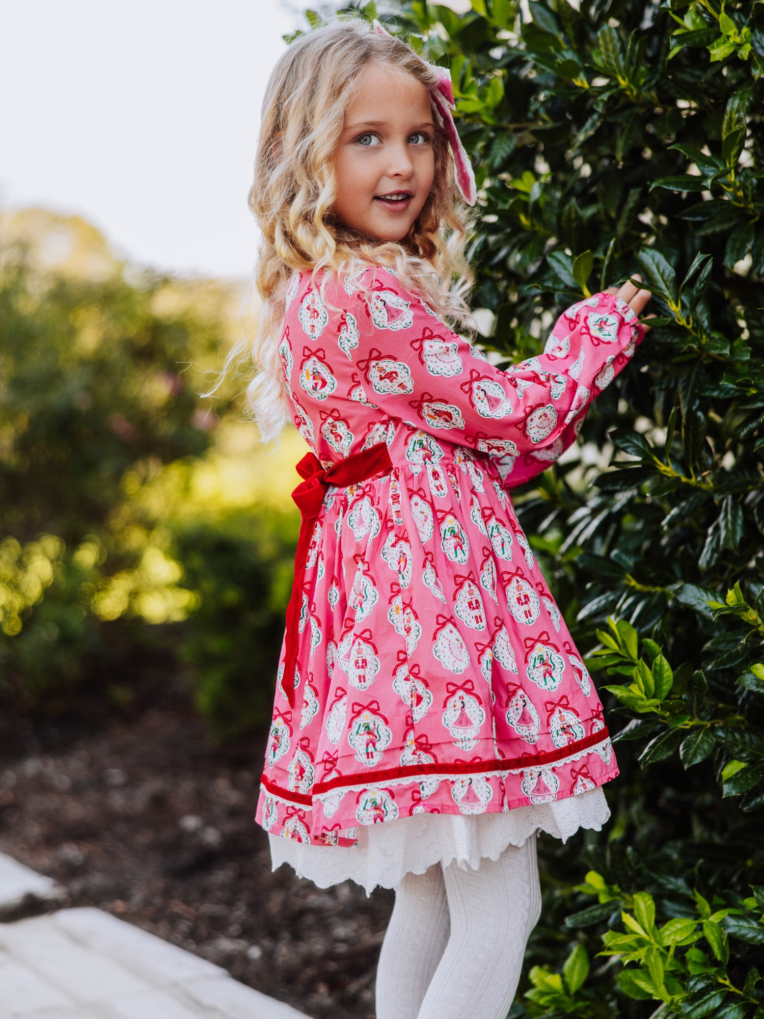 Girl standing outside next to greenery wearing a pink holiday themed dress.