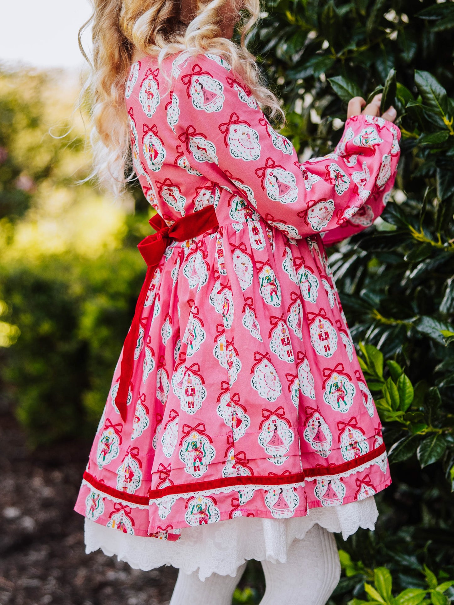 Girl standing outside next to greenery wearing a pink holiday themed dress.
