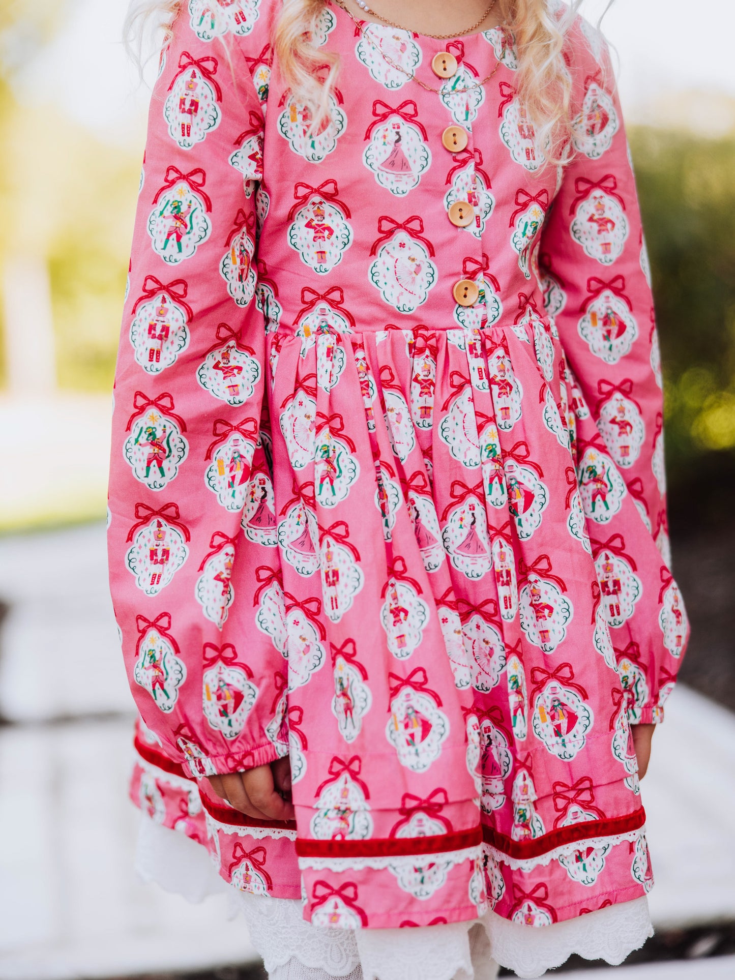Girl standing outside next to greenery wearing a pink holiday themed dress. Up close shot of buttons on bodice of dress. 