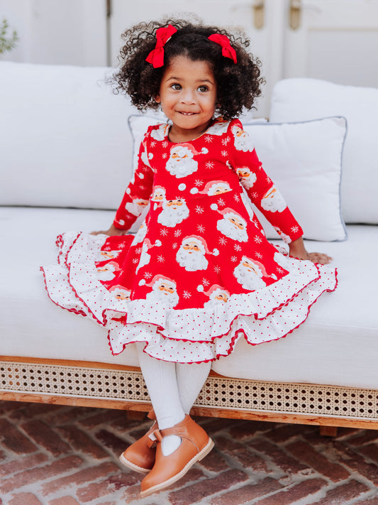 Girl sitting on couch wearing a red jolly santa printed dress with white tights and brown boots. 