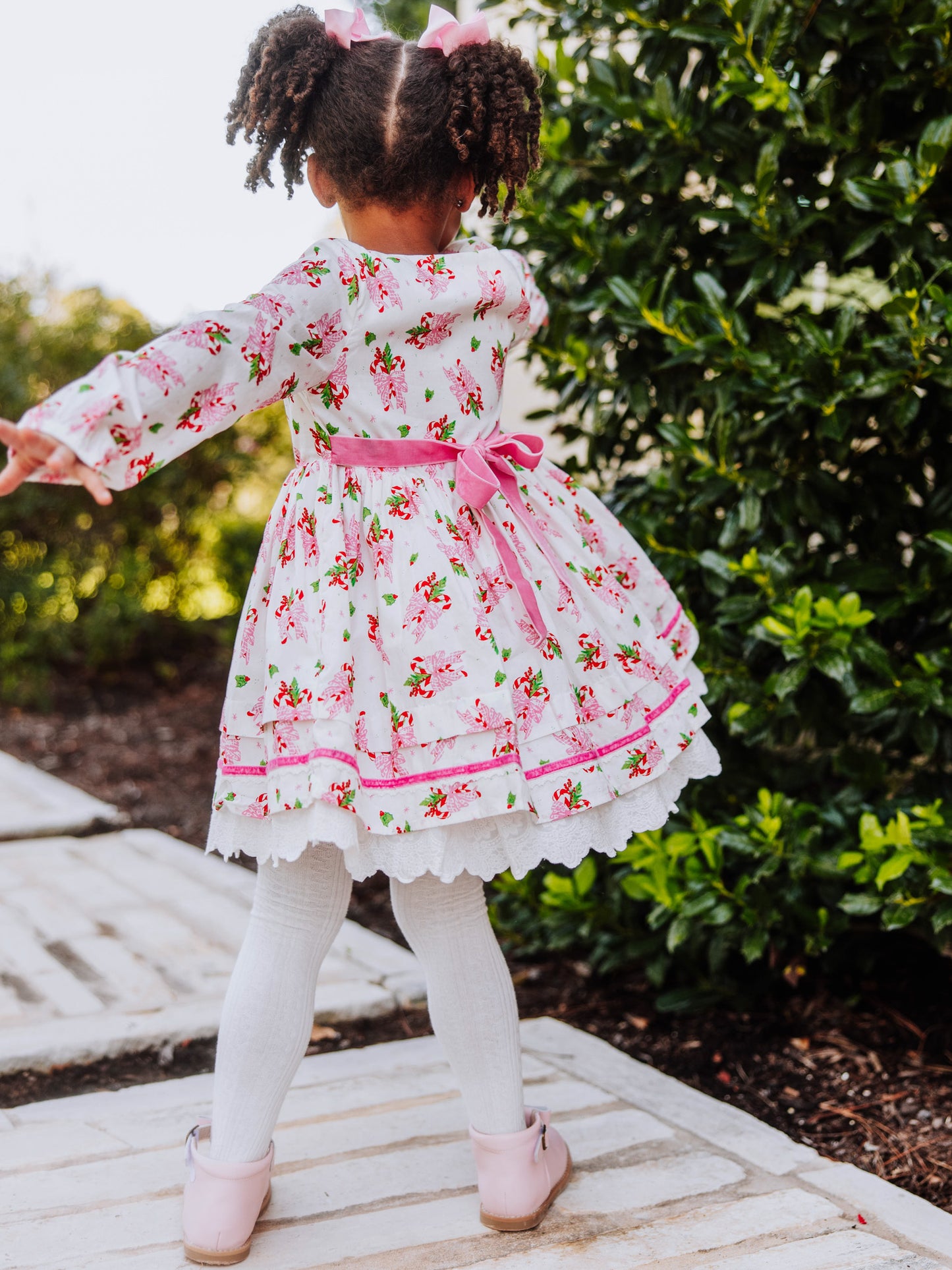 Girl standing outside next to greenery wearing a holiday themed dress and model is slightly spinning to show the pink velvet tie at back. 