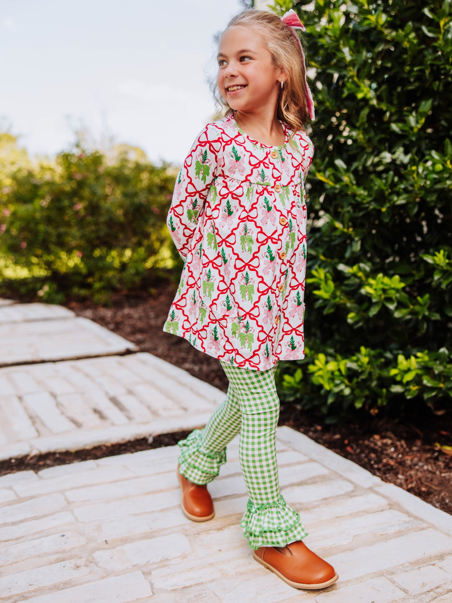 Girl has hands on her hips and is wearing a holiday bow themed top with green gingham bottoms with ruffles at ankles. 