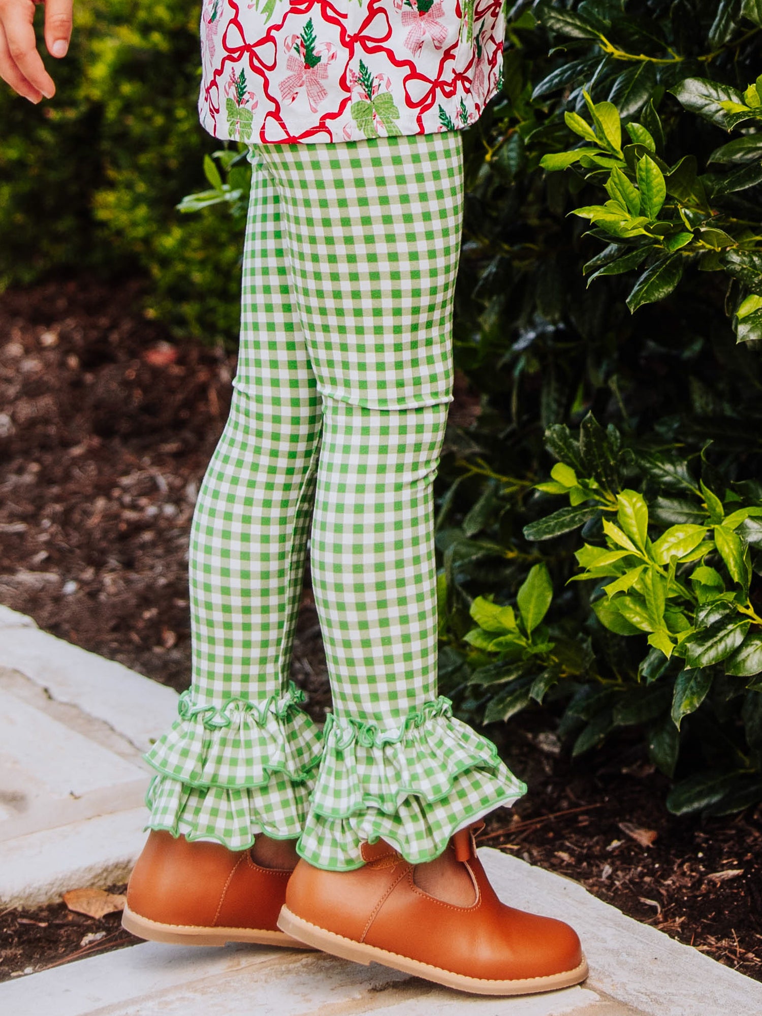 Girl has hands on her hips and is wearing a holiday bow themed top with green gingham bottoms with ruffles at ankles. 