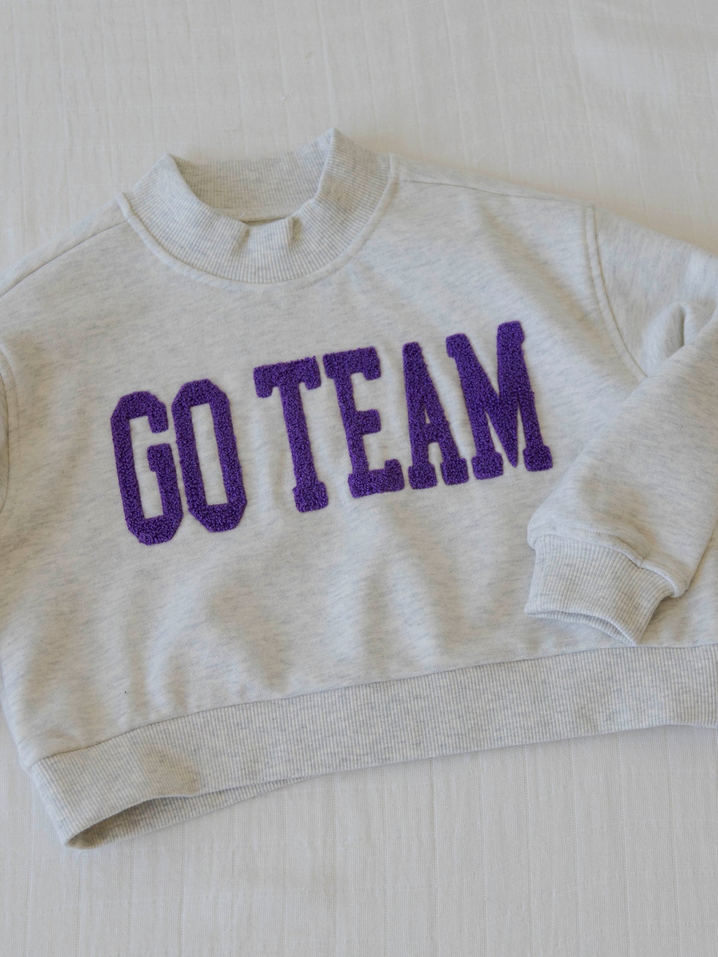 Flat lay of a heathered gray sweater that says GO TEAM in purple.