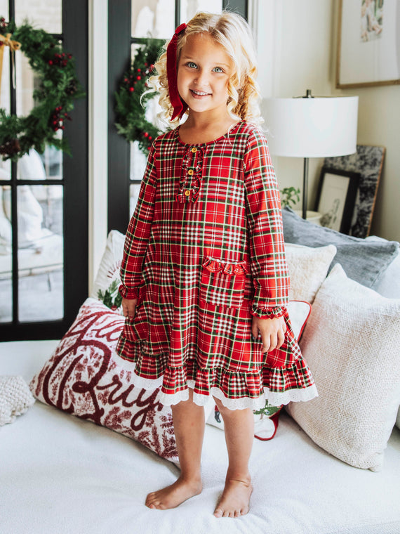 Girl standing on couch wearing play dress with red holiday plaid print.