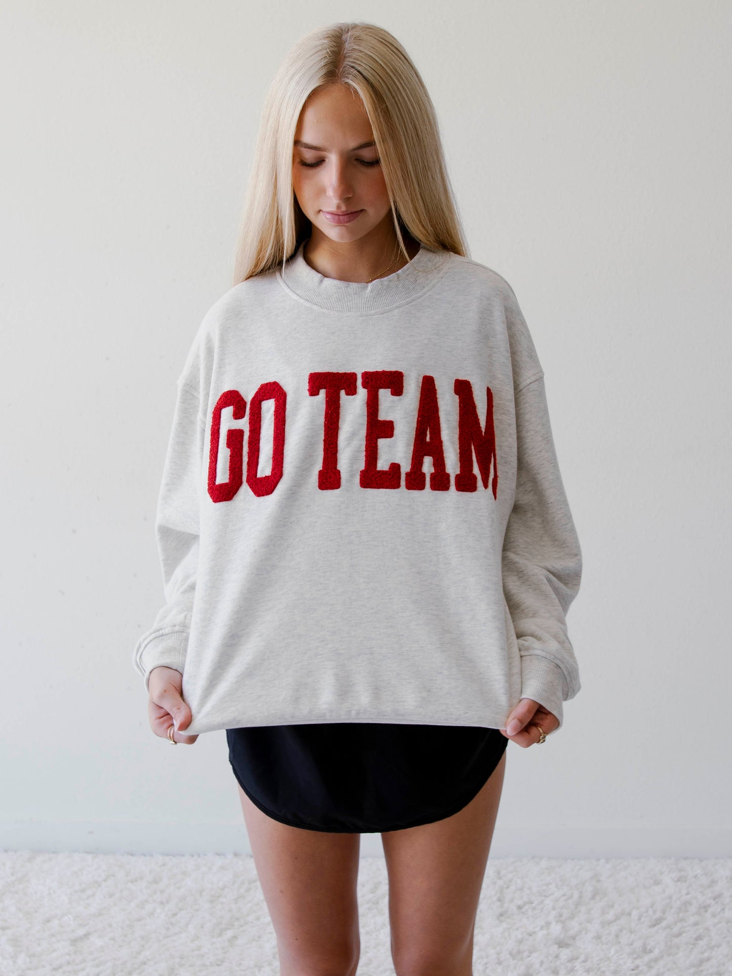 Girl in studio wearing a heathered gray sweatshirt that says GO TEAM  on the front in crimson red. 