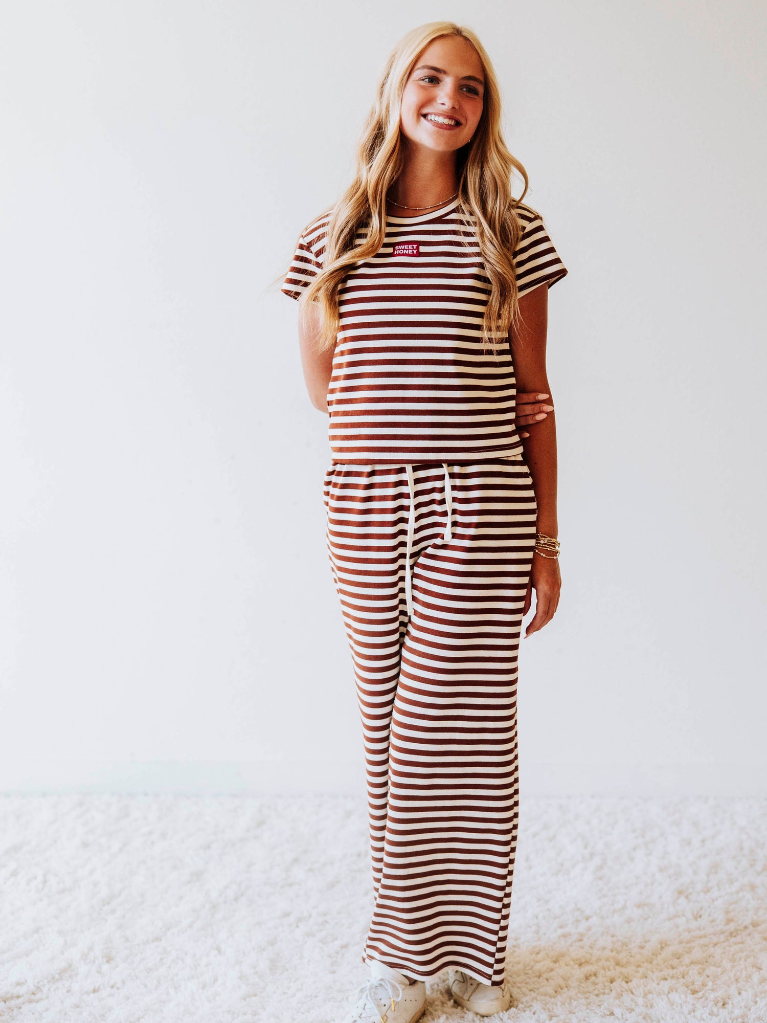 Model is wearing a chocolate and ivory striped, two piece pant set. Top has a SweetHoney logo tag stitched at chest