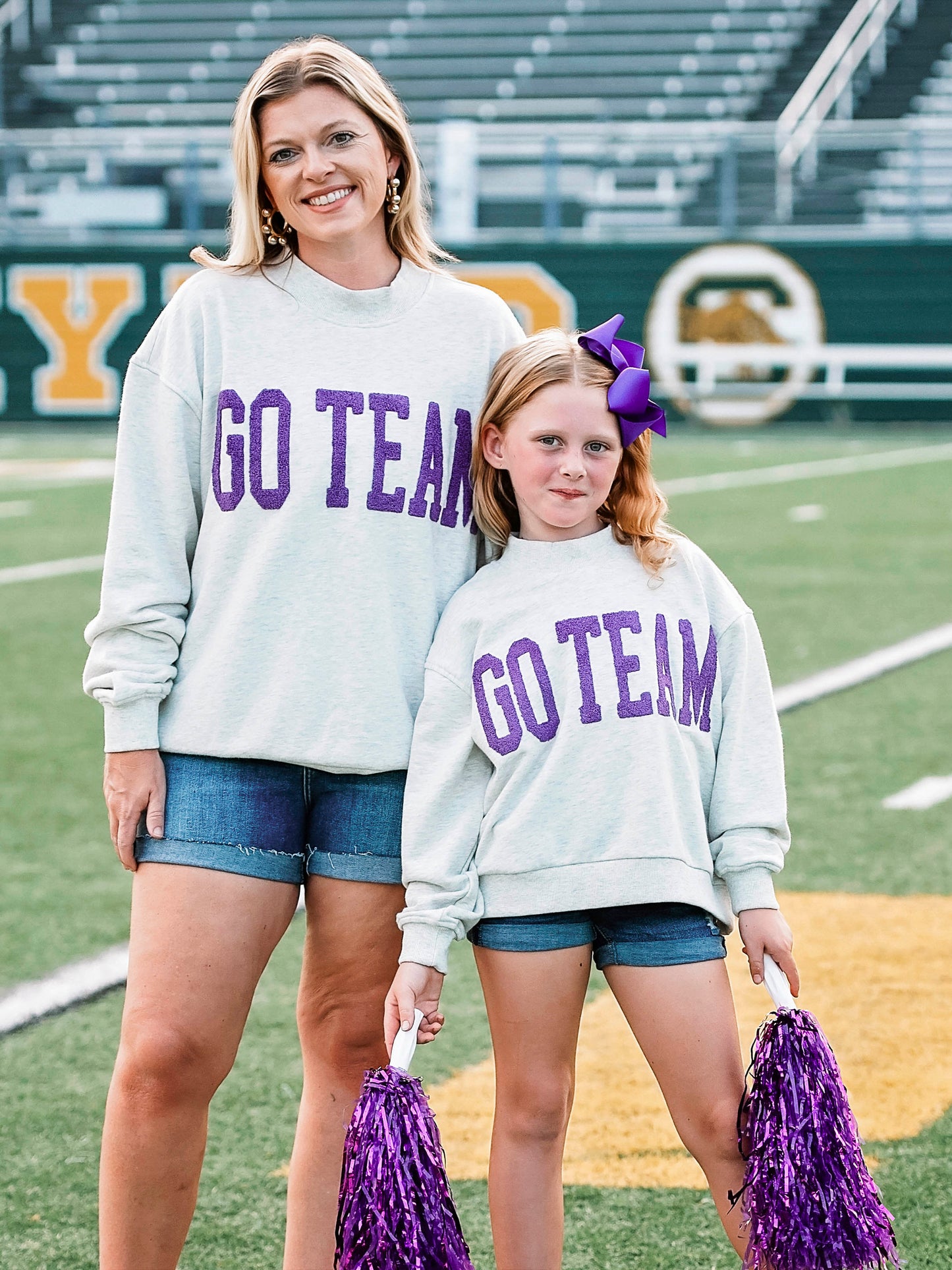 Women and child standing on a football field both wearing matching heathered gray sweatshirts that say GO TEAM on the front. Child is holding pom poms. 