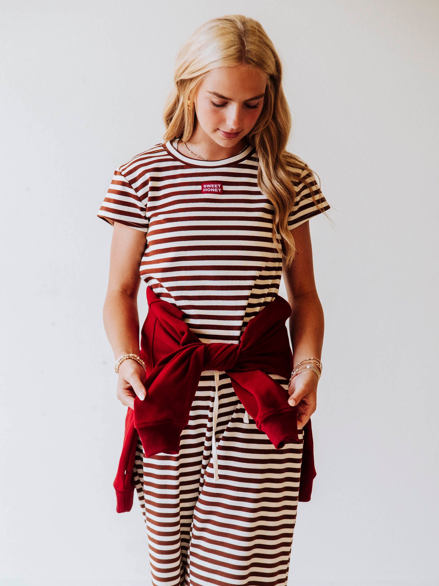 Model is wearing a chocolate and ivory striped pant set. Top has a stiched red and white Sweethoney tag centered at chest. Model also has a sweater tied around waist. 