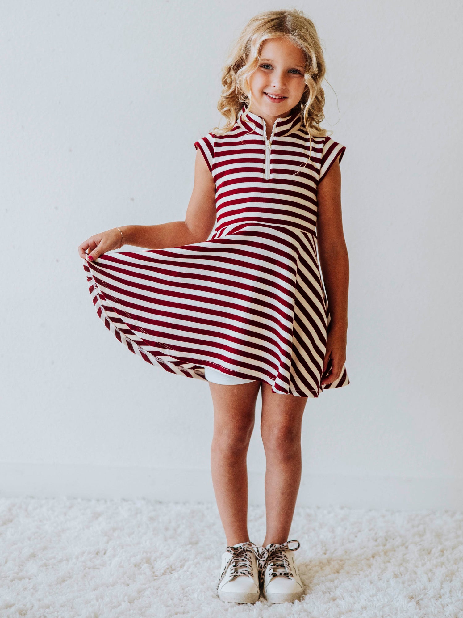 Model wearing crimson and ivory striped dress. Model is holding corner of dress and is wearing white shoes. 
