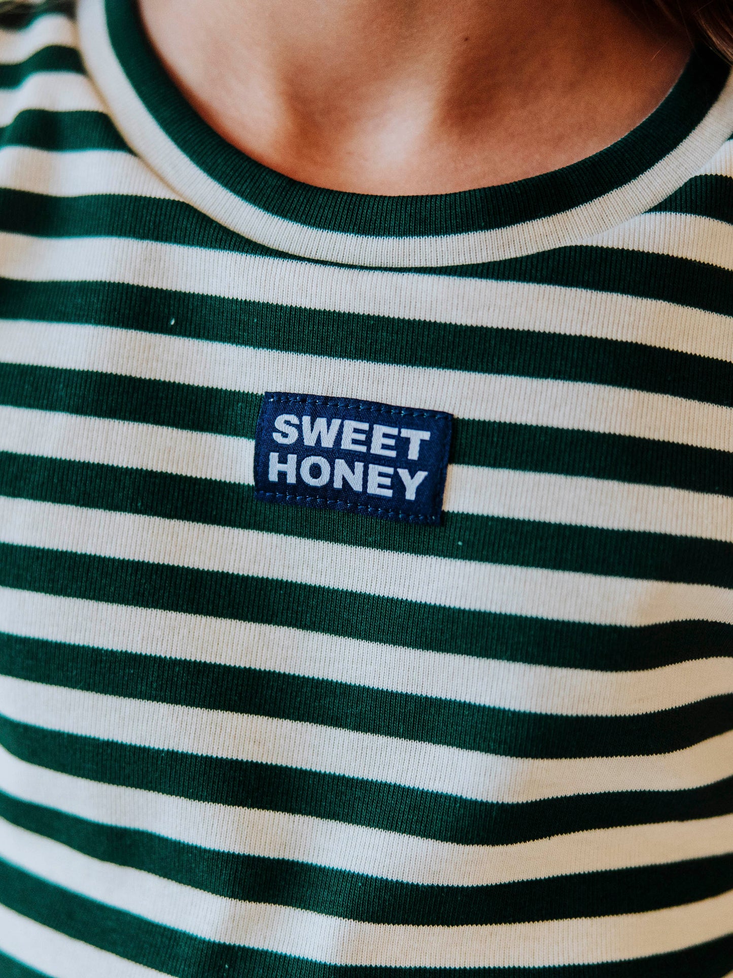 Up close picture of a green and ivory striped shirt with a SweetHoney navy and white logo tag in the center of chest. 
