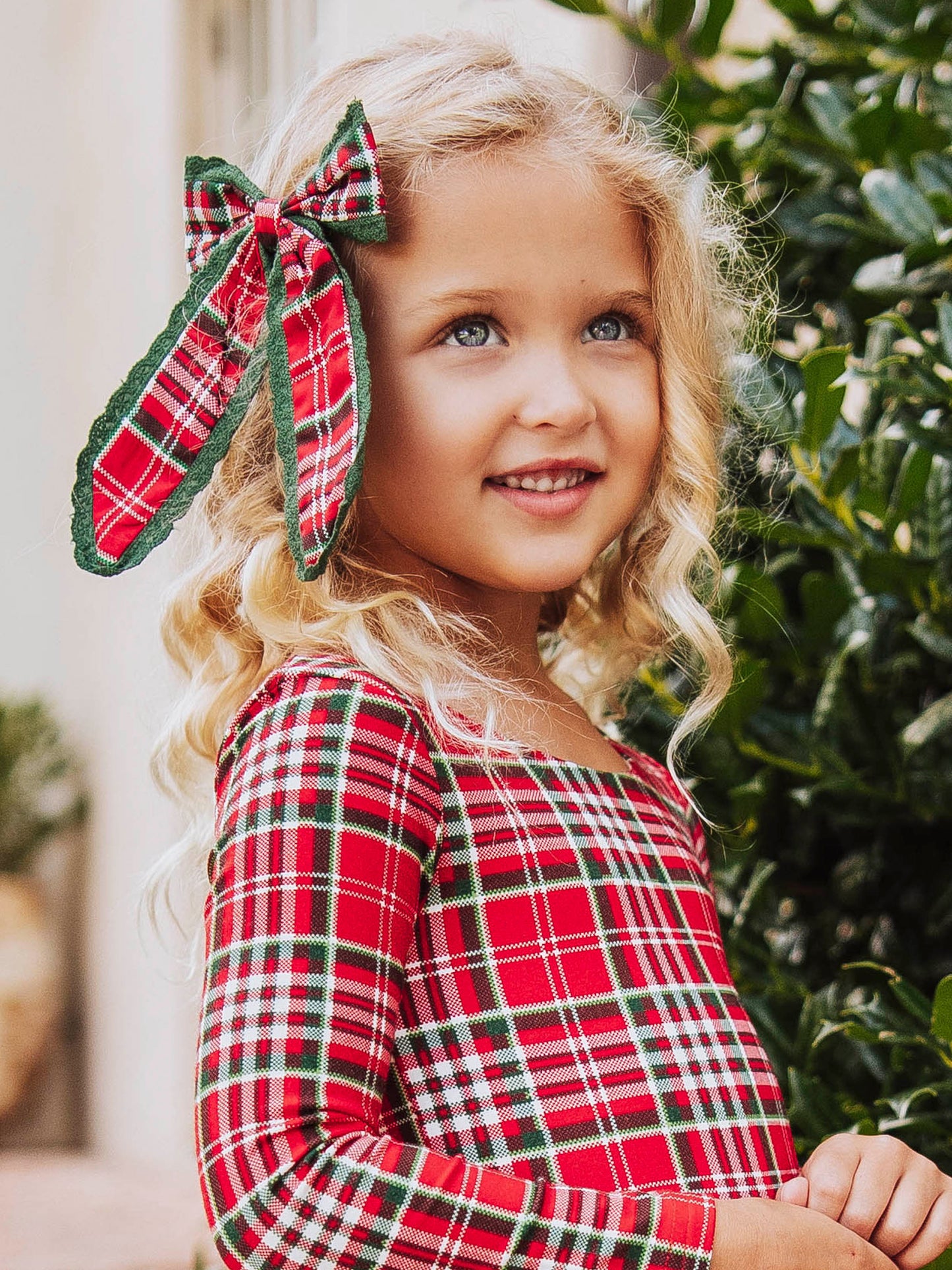 Up close shot of girl wearing a red traditional colored plaid bow on the side of her head. Model wearing the same colored leggy romper too.