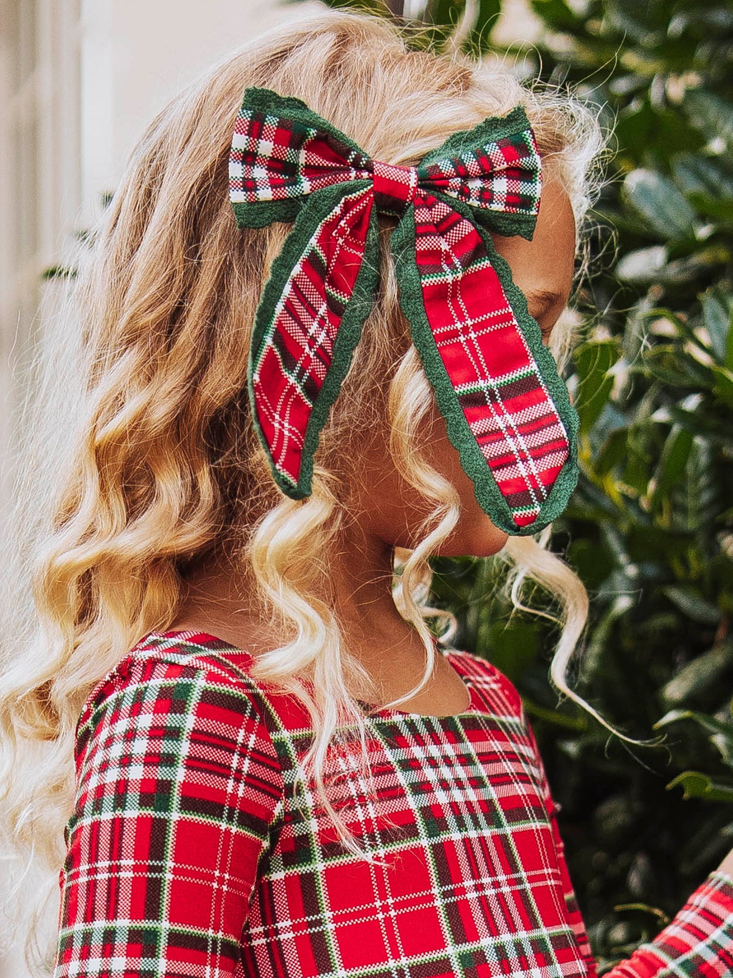 Up close shot of girl wearing a red traditional colored plaid bow on the side of her head. Model wearing the same colored leggy romper too.