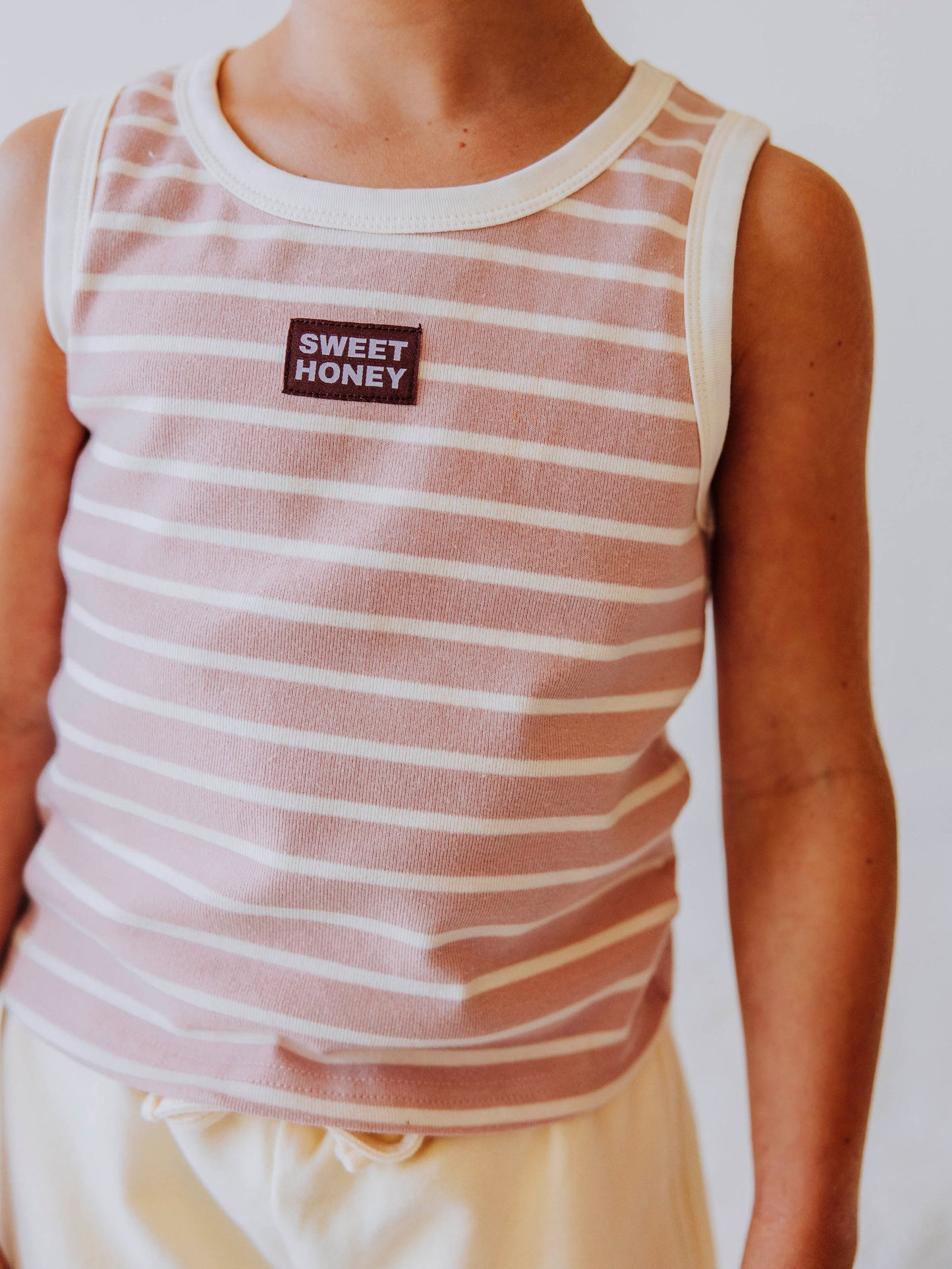Model is wearing a pink and ivory sleeveless shirt that has a sweethoney tag stitched in the middle of shirt on chest. model is wearing cream colored shorts. 