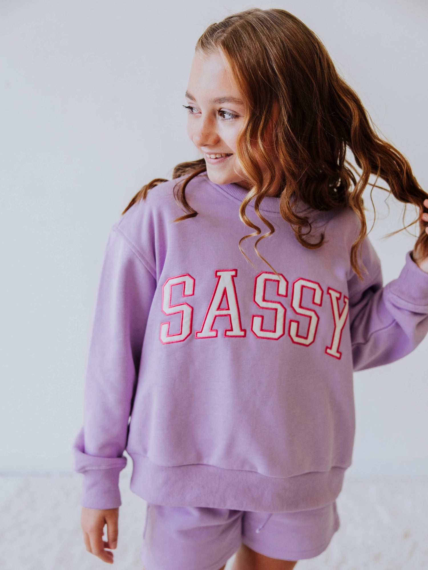Model is wearing a two piece, purple in color sweat set. the top has SASSY on the front in white letters that are outlined in pink. 