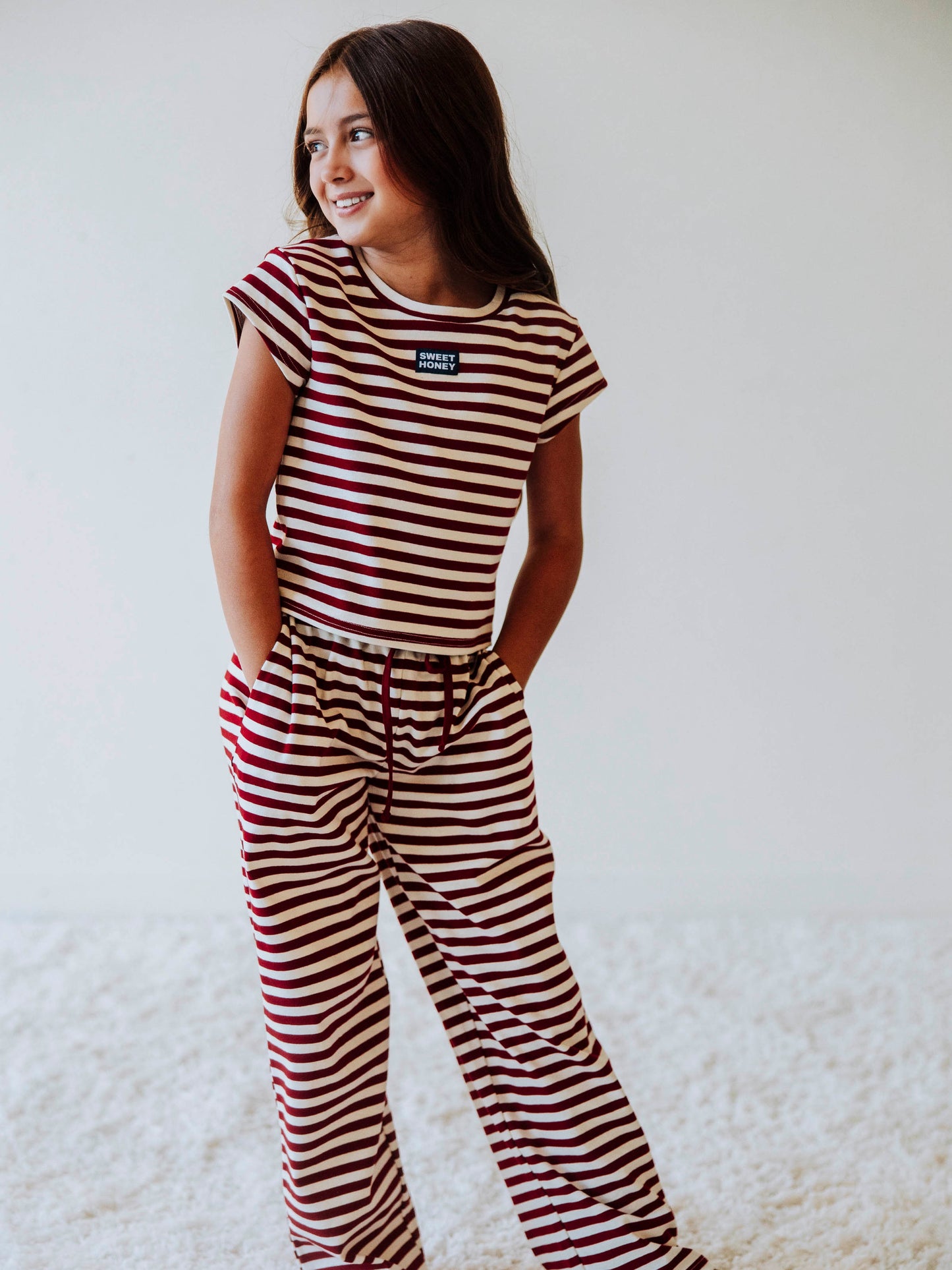 Girl is wearing a two-piece red and ivory striped pant set. Girl is wearing tan clogs and is posing. Girls hands are in her pocket. 