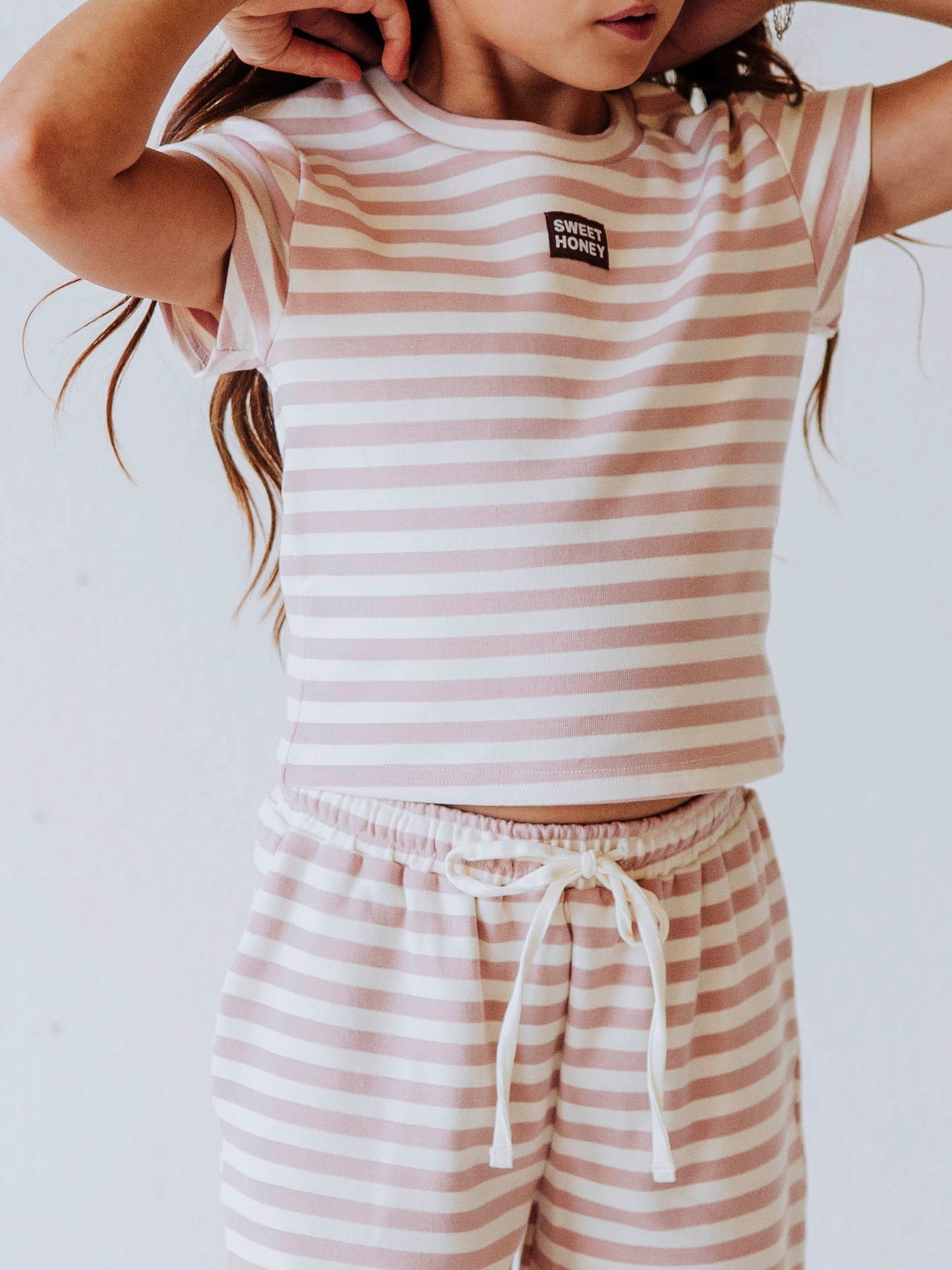 Girl is wearing a two-piece, pink and ivory striped pant set. There is a SweetHoney logo tag stitched on the center of shirt. 
