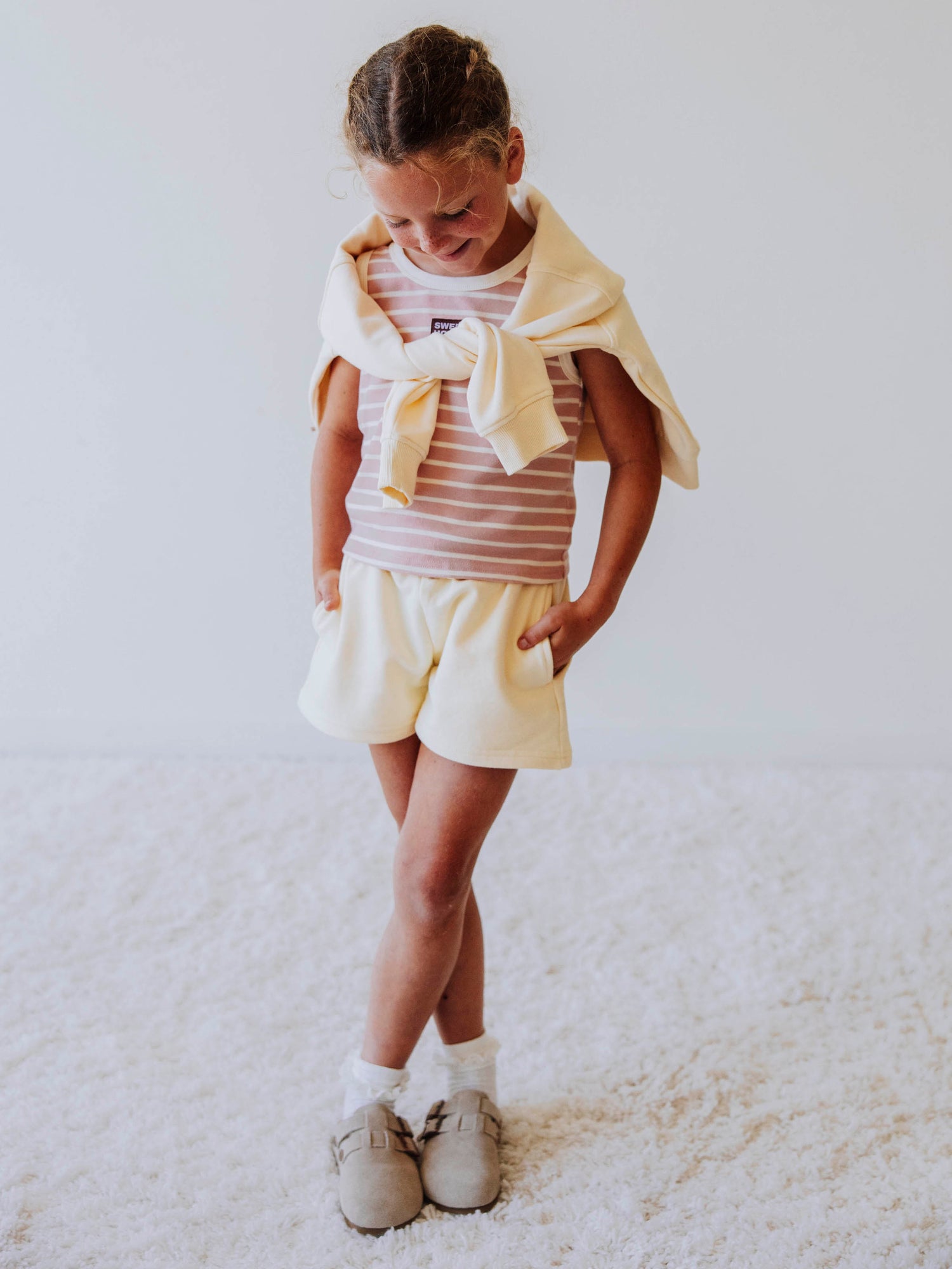 Model is wearing an ivory and pink sleeveless shirt, an ivory colored sweater is draped over shoulders, model is wearing ivory shorts with clog shoes. 