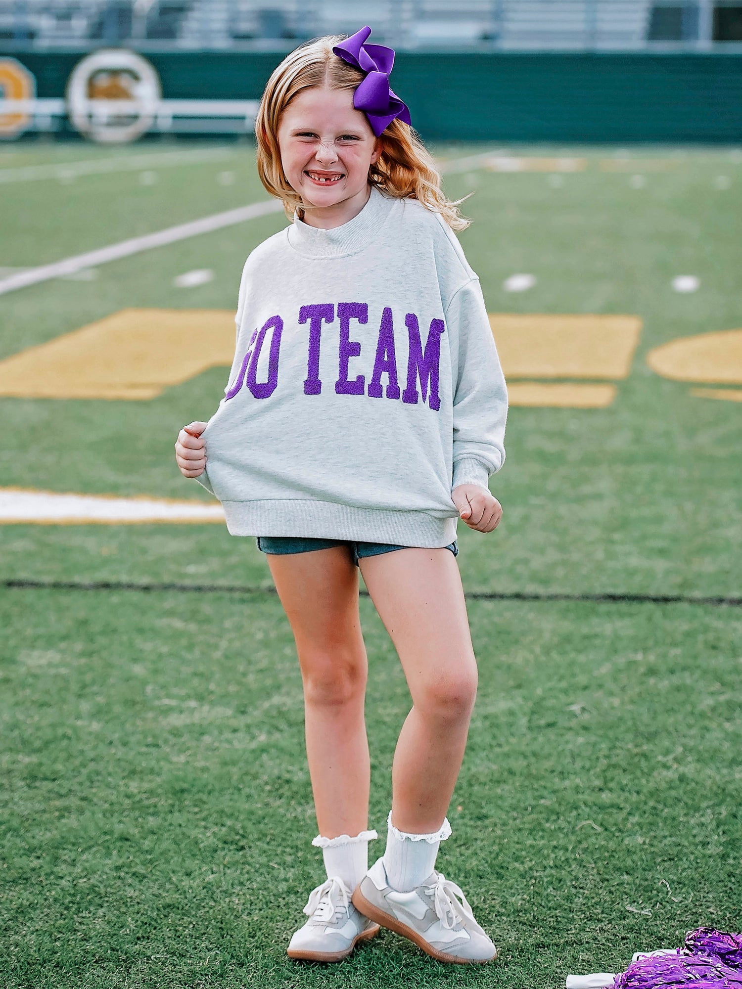 Girl wearing a heathered gray sweatshirt that says GO TEAM on the front in purple. Girl is standing on a football field. 