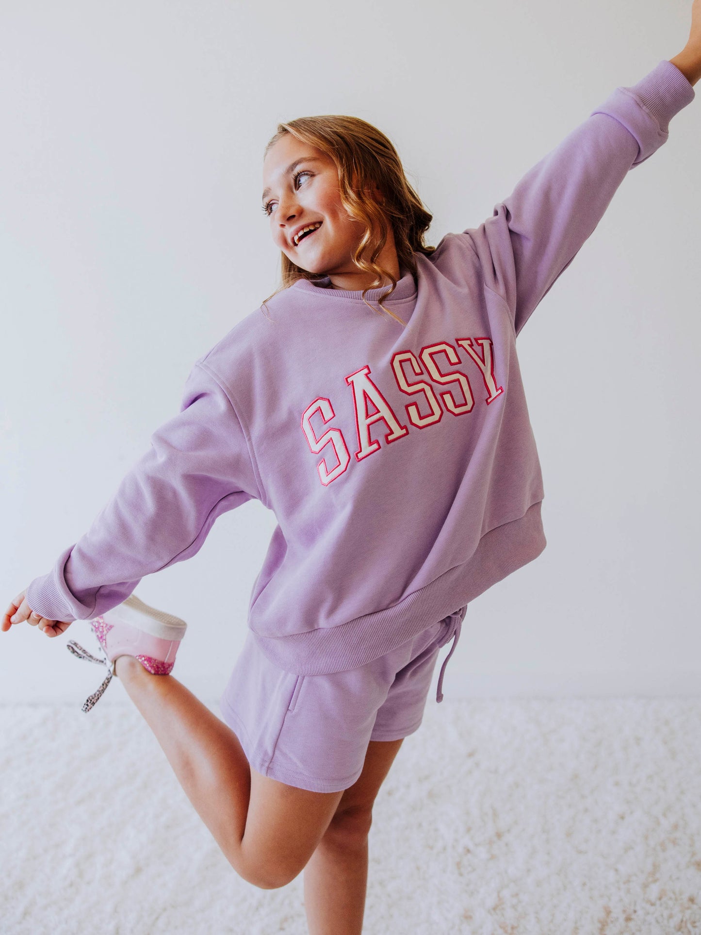 Model is wearing a two piece, purple in color sweat set. the top has SASSY on the front in white letters that are outlined in pink. 