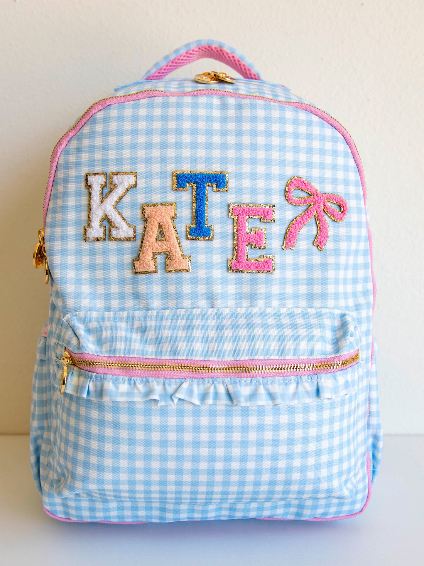 Blue gingham backpack with light pink zippers and gold zipper pulls and different color letter patches