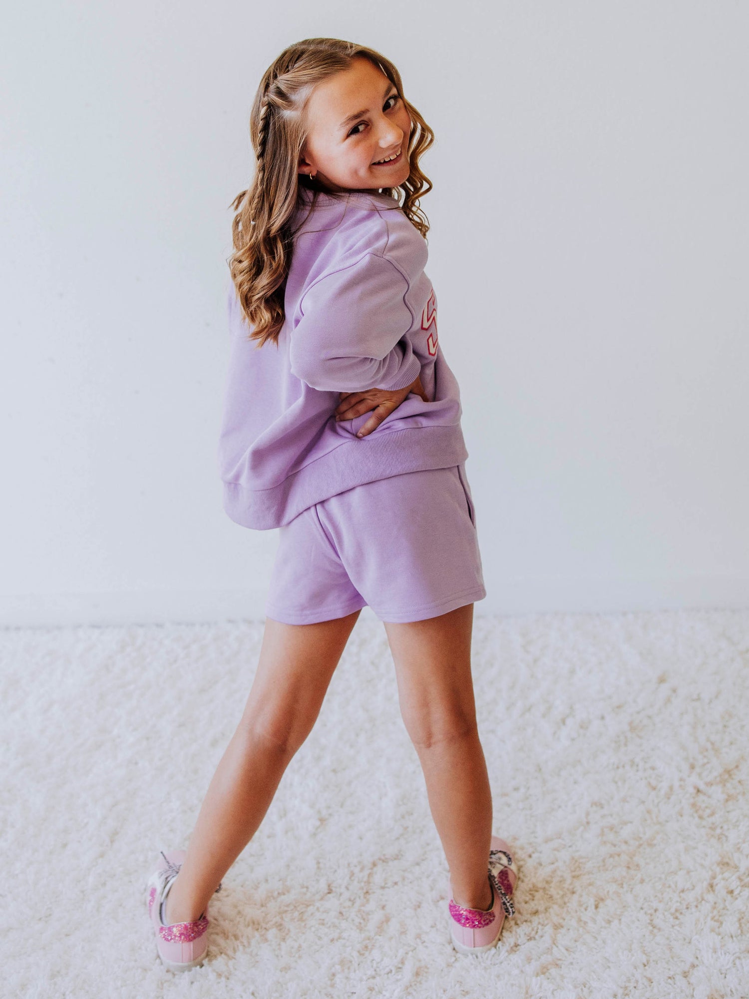 Model is wearing a two piece, purple in color sweat set. the top has SASSY on the front in white letters that are outlined in pink. Picture shows model from behind. 