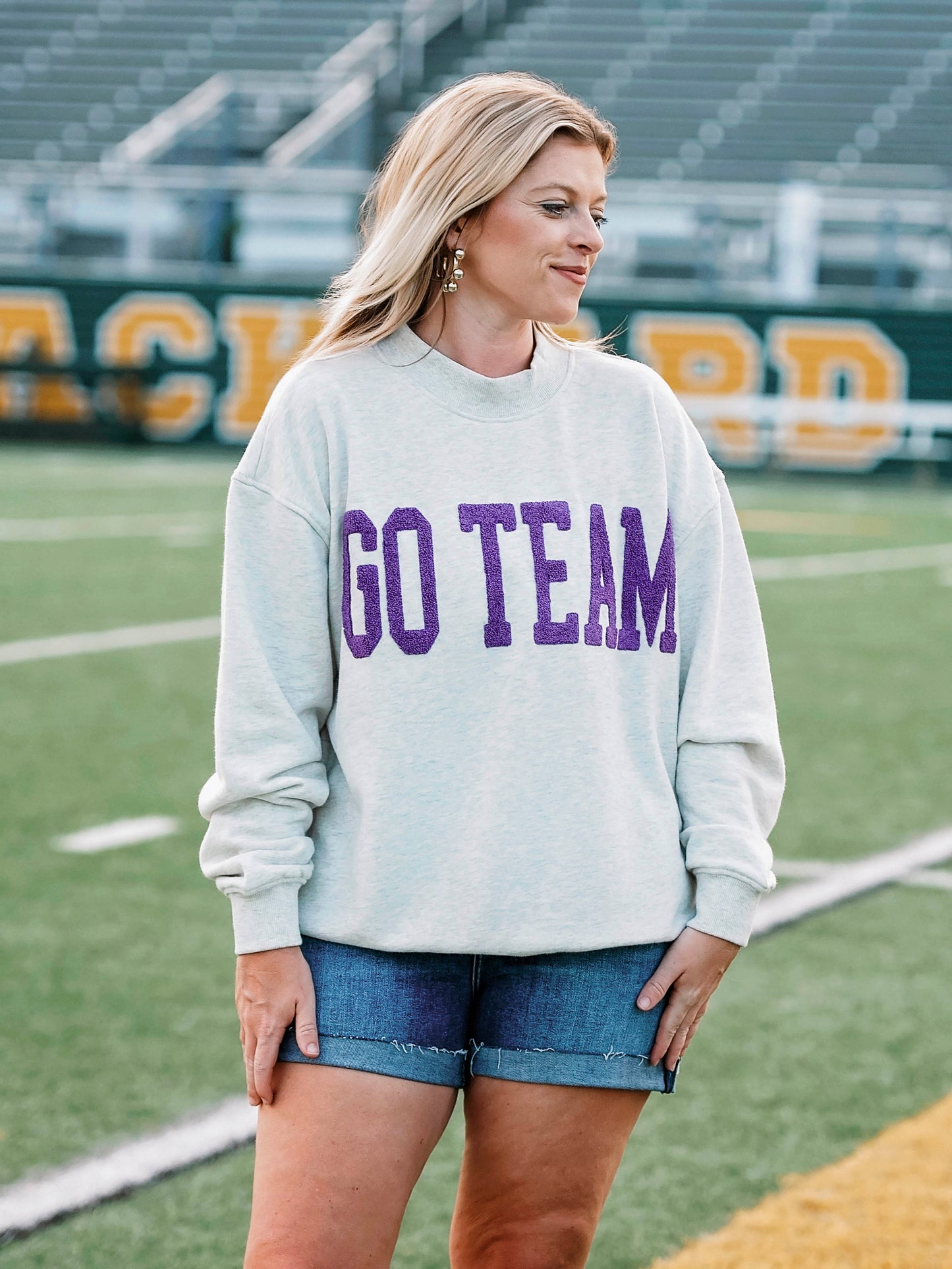 Women at a football field wearing a heathered gray sweatshirt that says GO TEAM on the front in purple. 