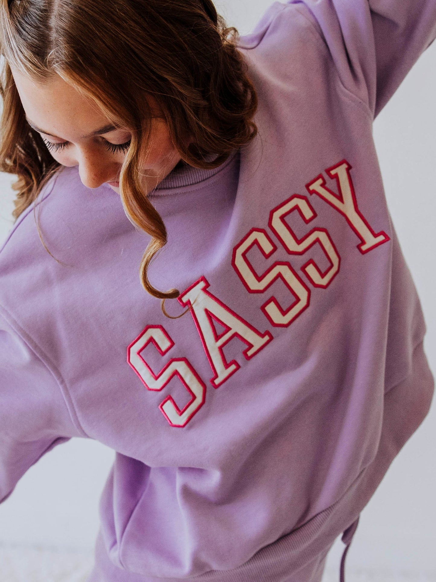 Model is wearing a two piece, purple in color sweat set. the top has SASSY on the front in white letters that are outlined in pink. Picture is a close up of top. 