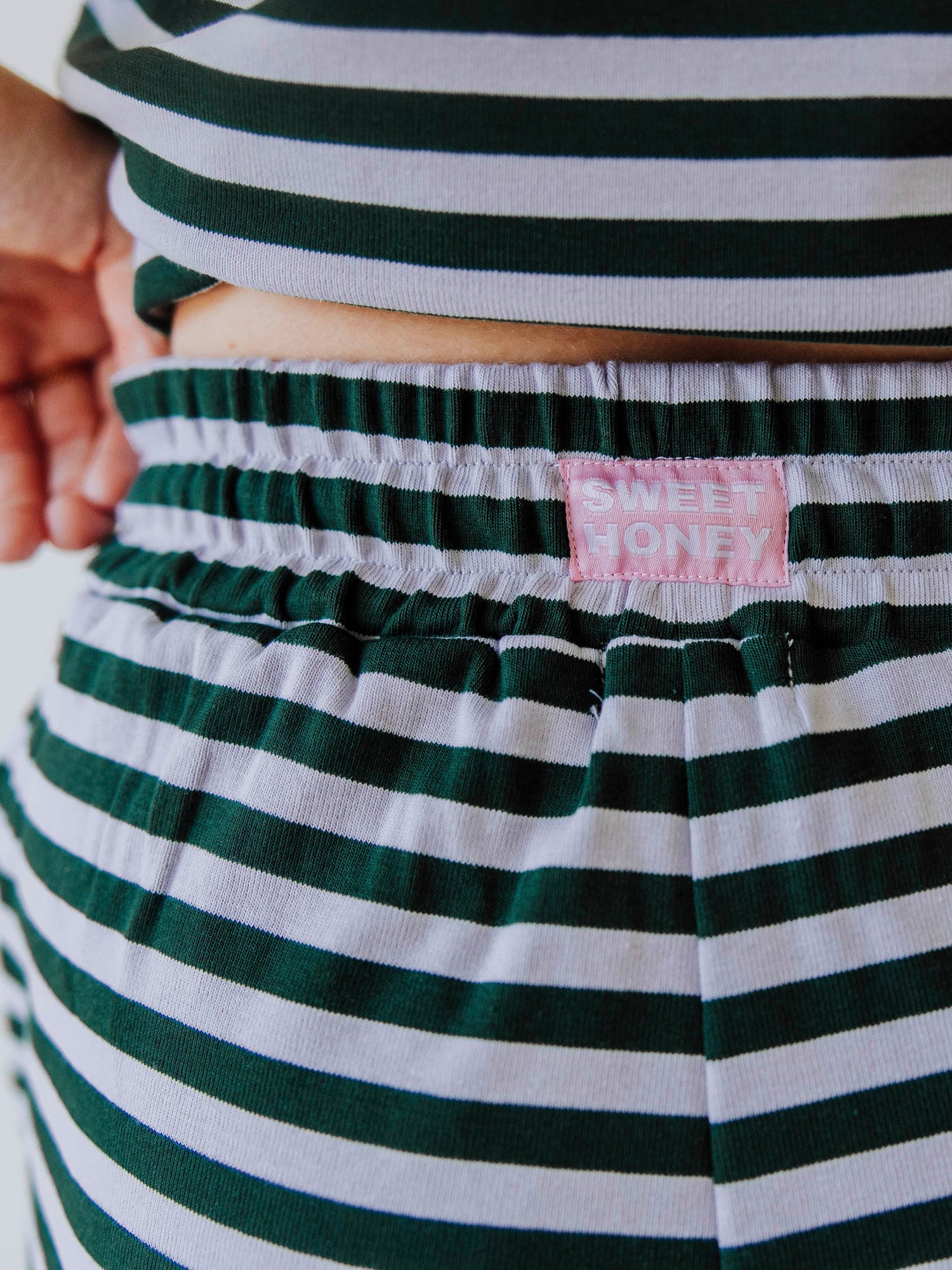 Model is wearing a green and lavender striped two-piece set with a green sweater draped over her shoulder. Up close picture of the SweetHoney logo tag on the back of waistband.