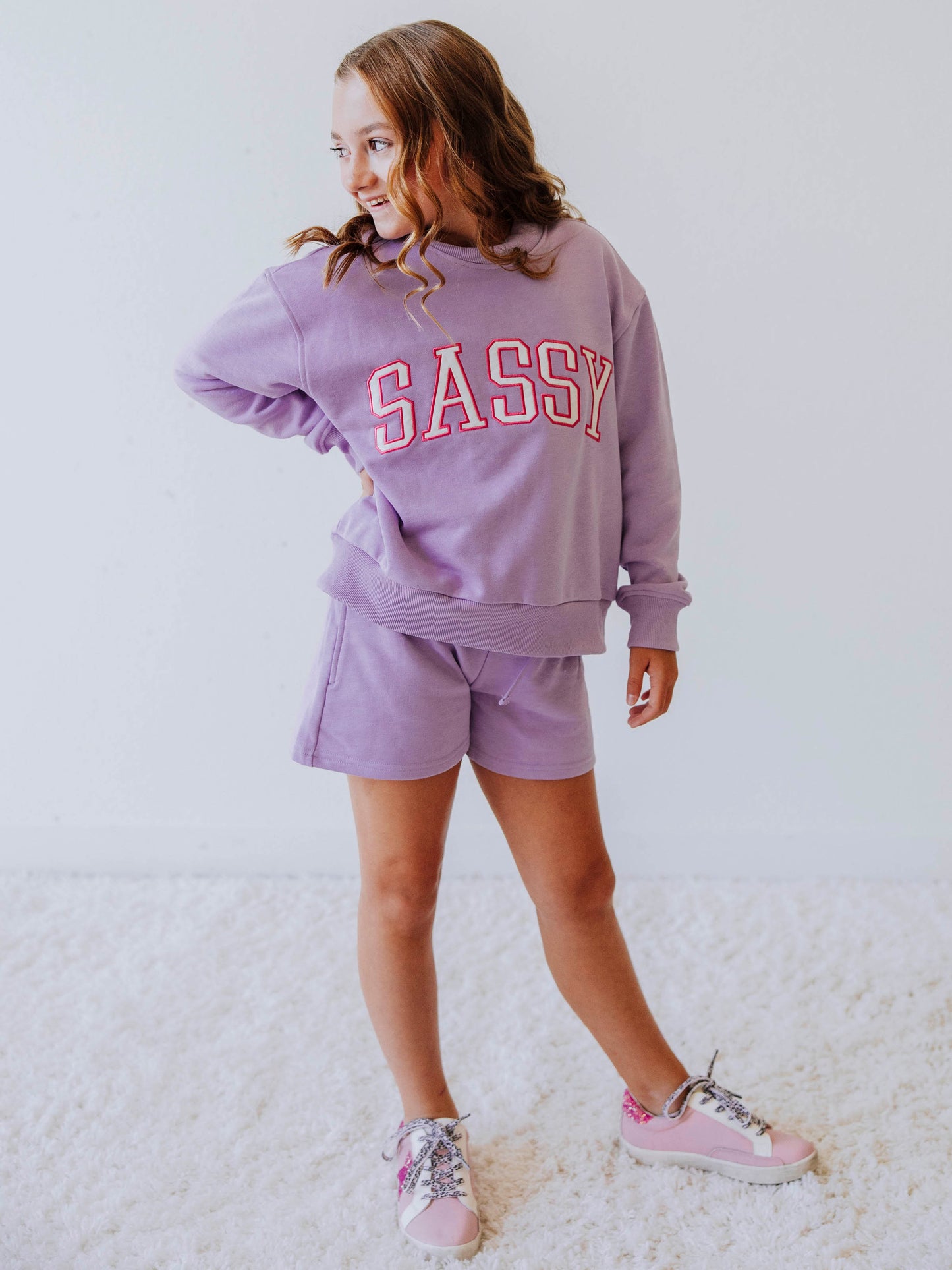 Model is wearing a two piece, purple in color sweat set. the top has SASSY on the front in white letters that are outlined in pink. 