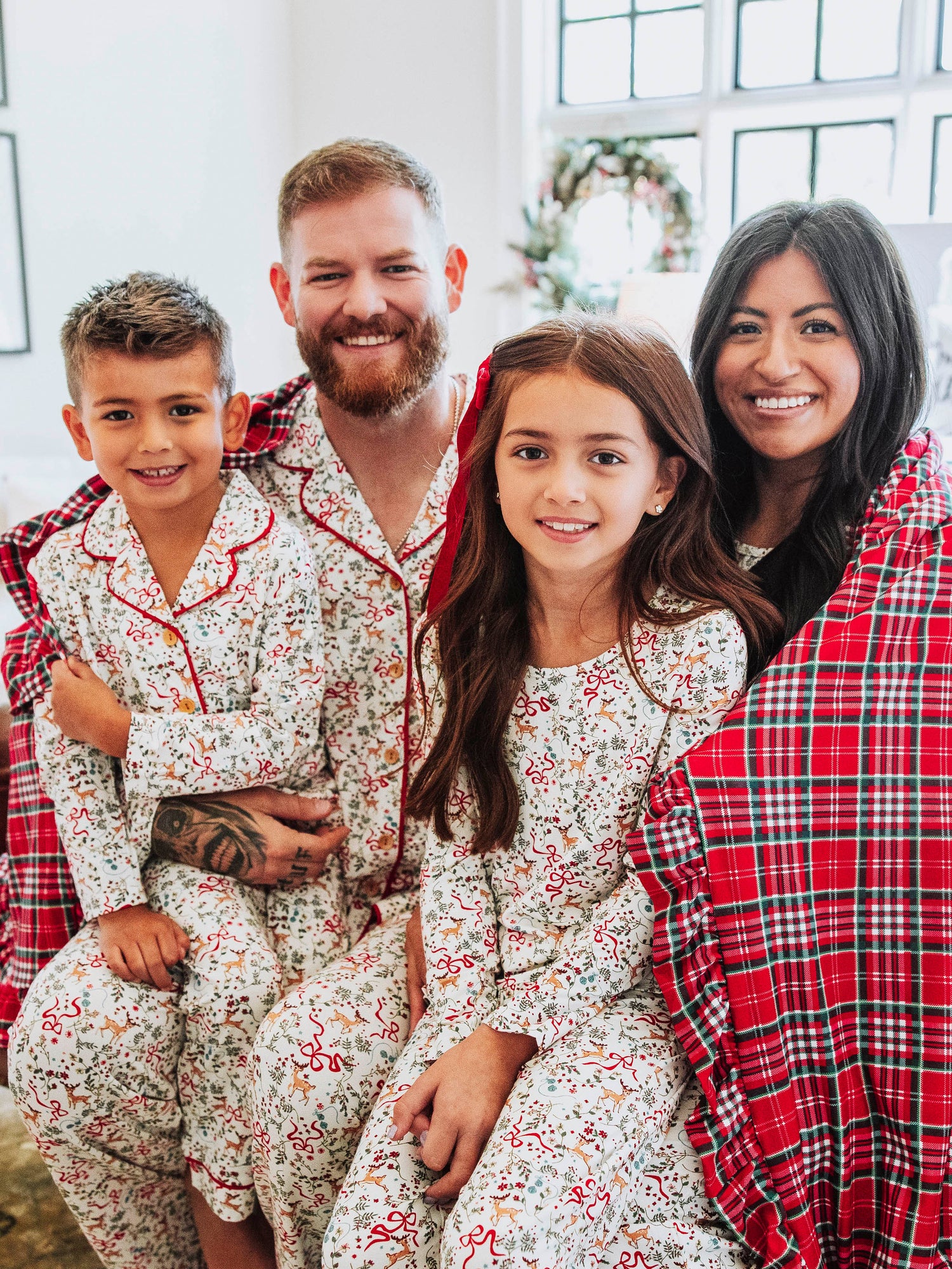 A family of four all wearing Christmas themed pajamas and wrapped up in a traditional plaid Christmas printed blanket. 