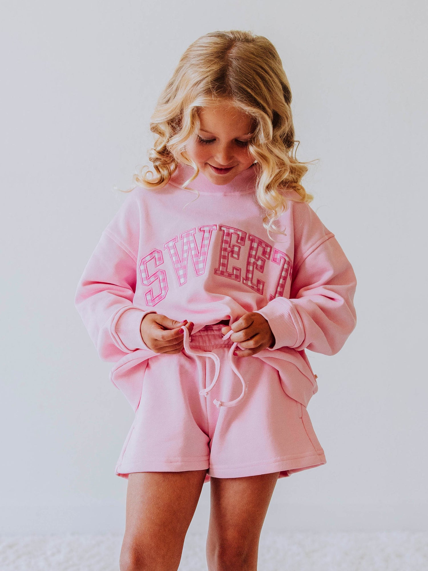 Girl is wearing a two-piece pink jogging set. The top says SWEET on the front and the letters have pink gingham checks in each. Bottoms are the same pink as top.